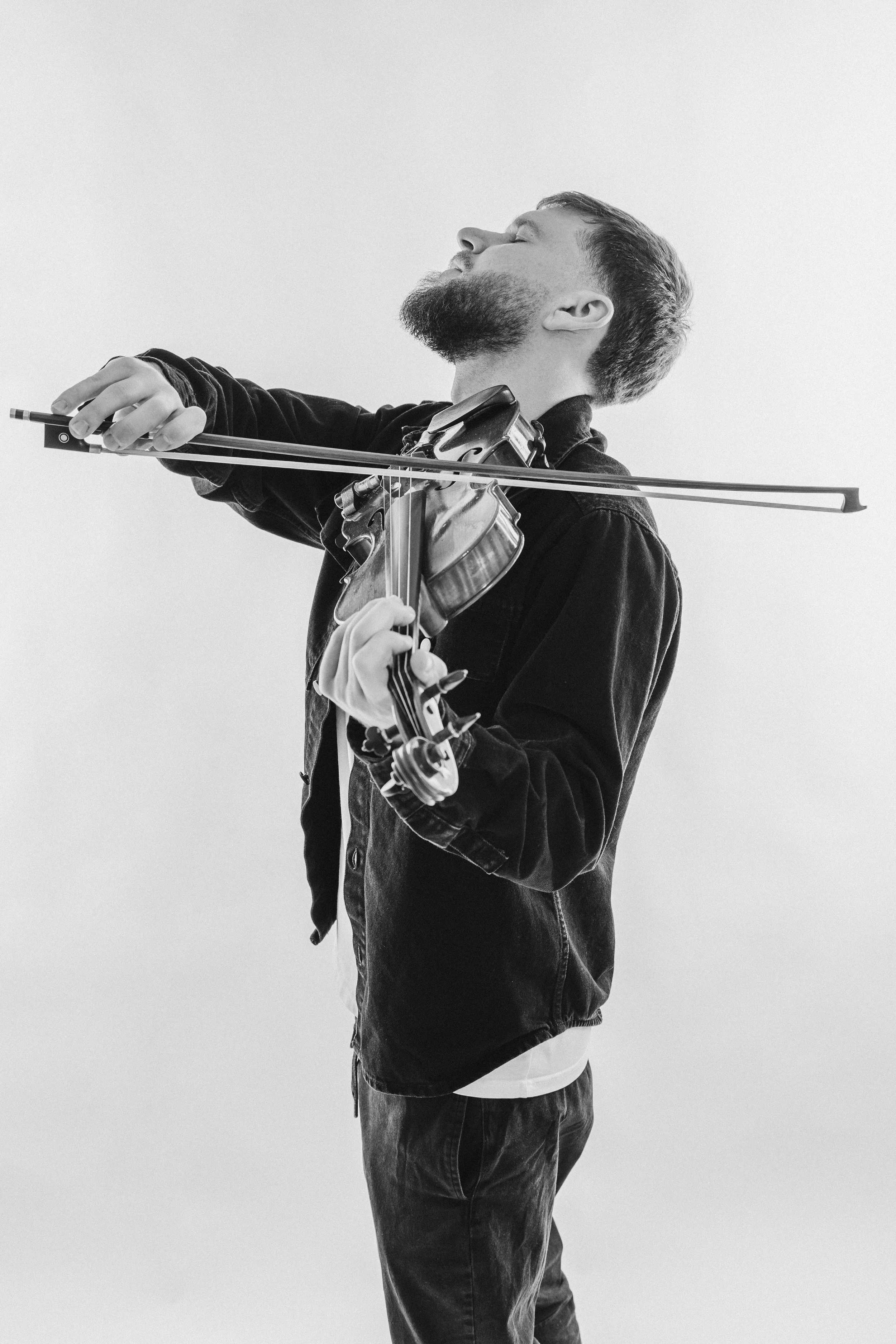 A man with a beard playing the violin, eyes closed, wearing a dark jacket, with a light background.