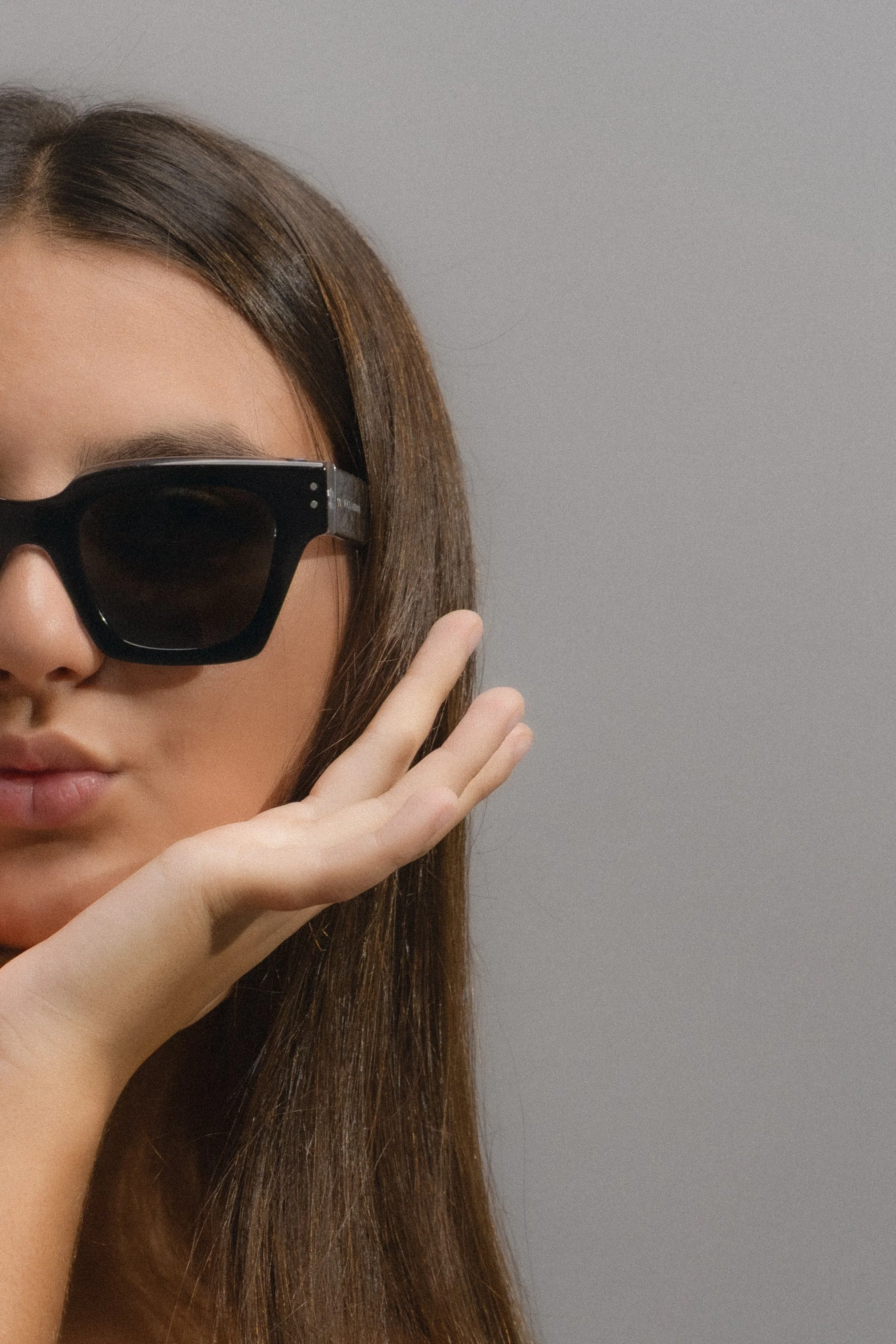 Close-up of a woman with long brown hair wearing black sunglasses touching her face against a plain gray background.