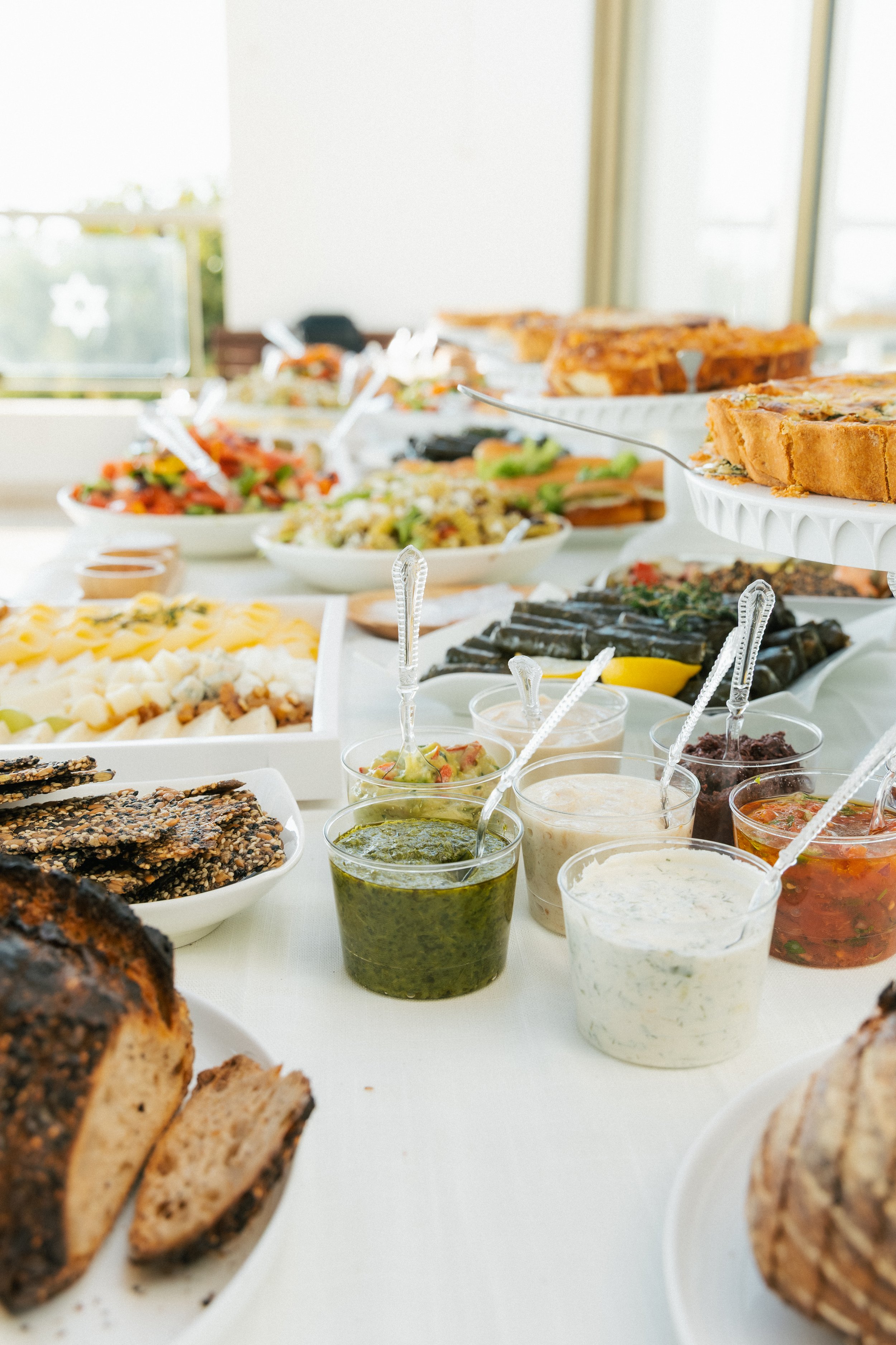 Assorted appetizers, salads, bread, spreads, and sauces at a buffet table in a well-lit room with large windows.