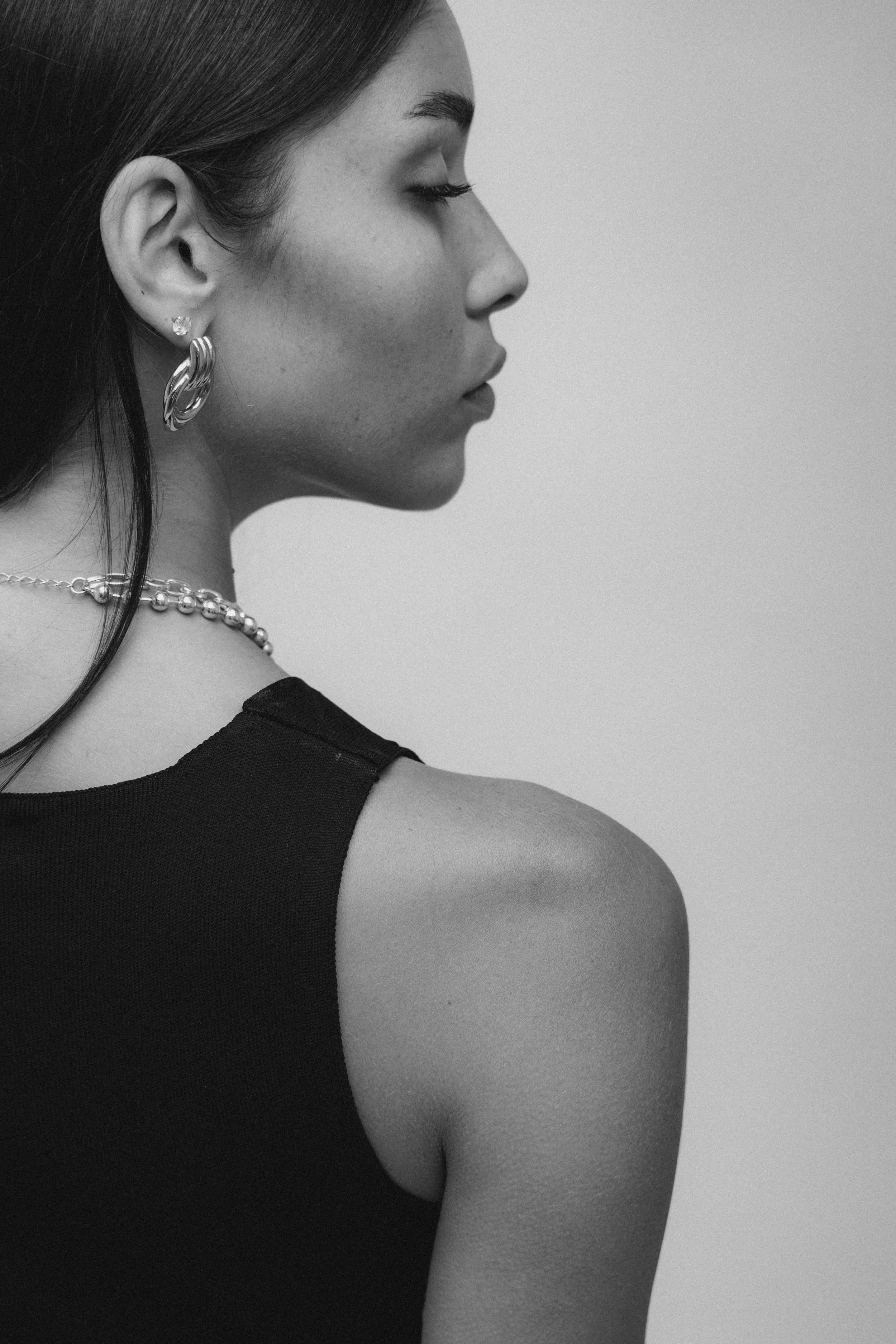 A close-up profile shot of a woman with smooth skin, wearing a black sleeveless top, layered jewelry including earrings, a necklace, and a chain on her shoulder. The image is in black and white.