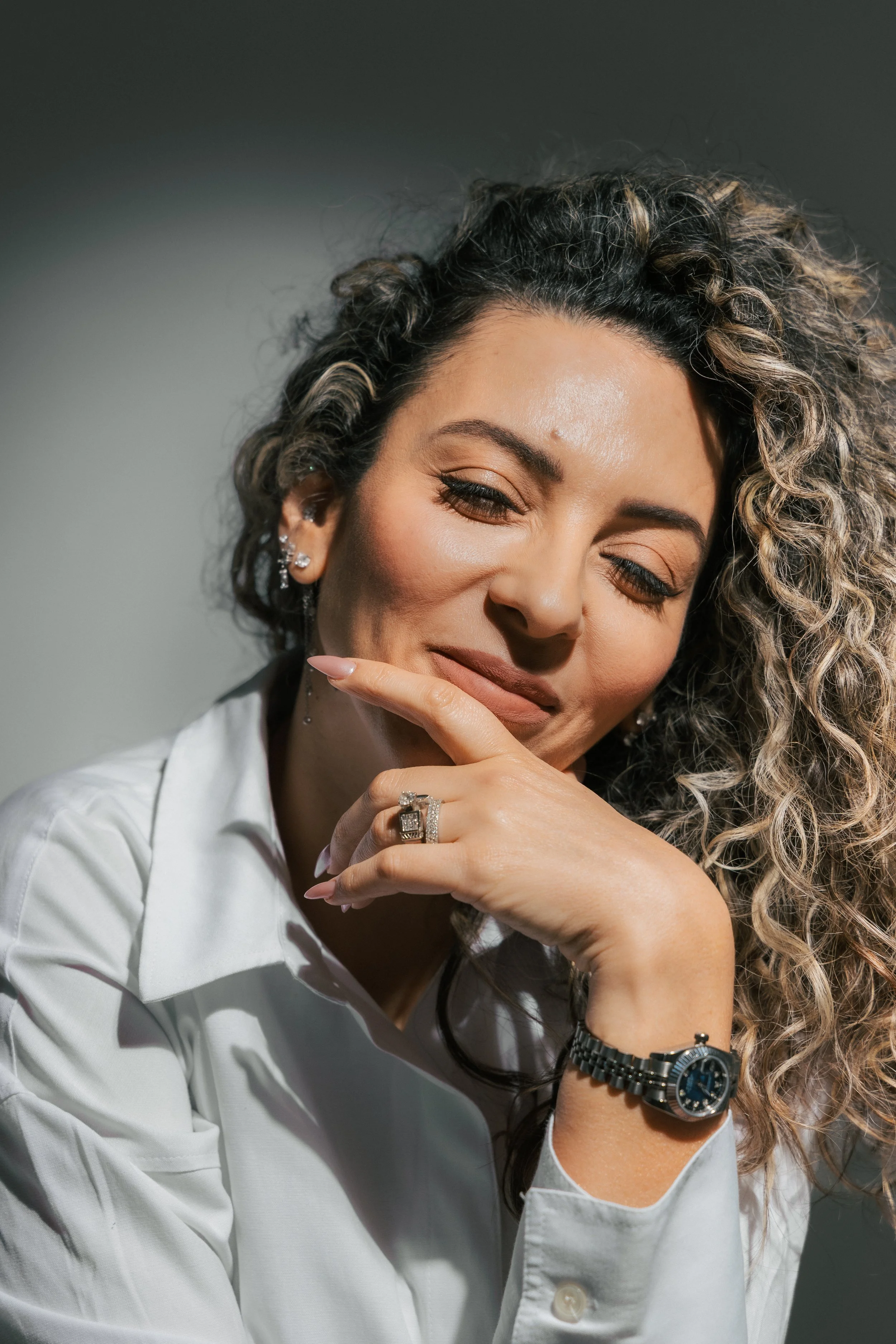 A woman with curly, blonde-highlighted hair, wearing a white shirt, jewelry, and a watch, smiling with her eyes closed and her hand near her face.
