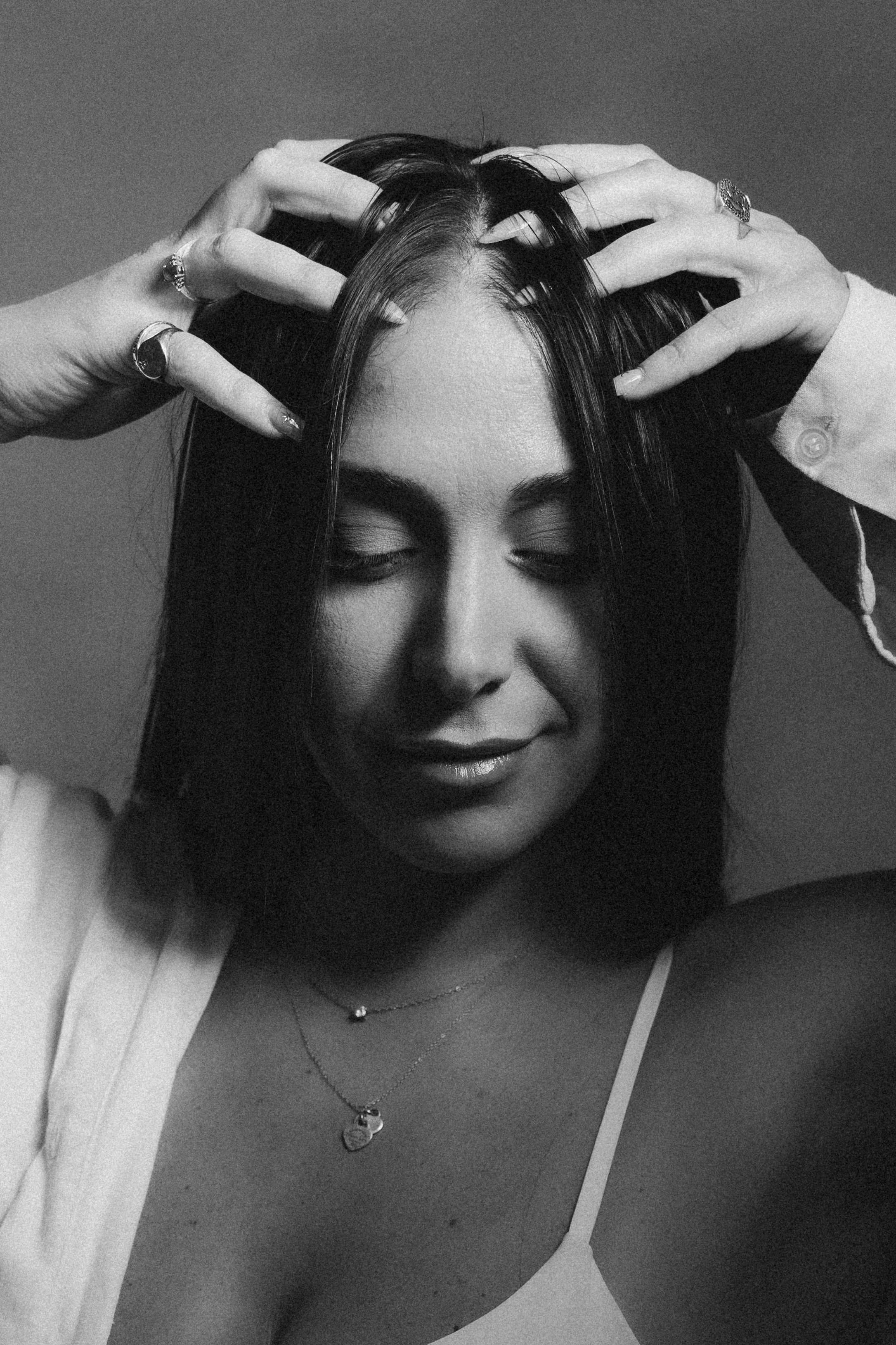 A woman with closed eyes, dark hair, and wearing jewelry, including rings and necklaces, is holding her hands on her head, in a close-up Black and White portrait. פורטריט נשיות, פורטריט בסטודיו, צילומי נשיות, עריכה אדיטוריאלית