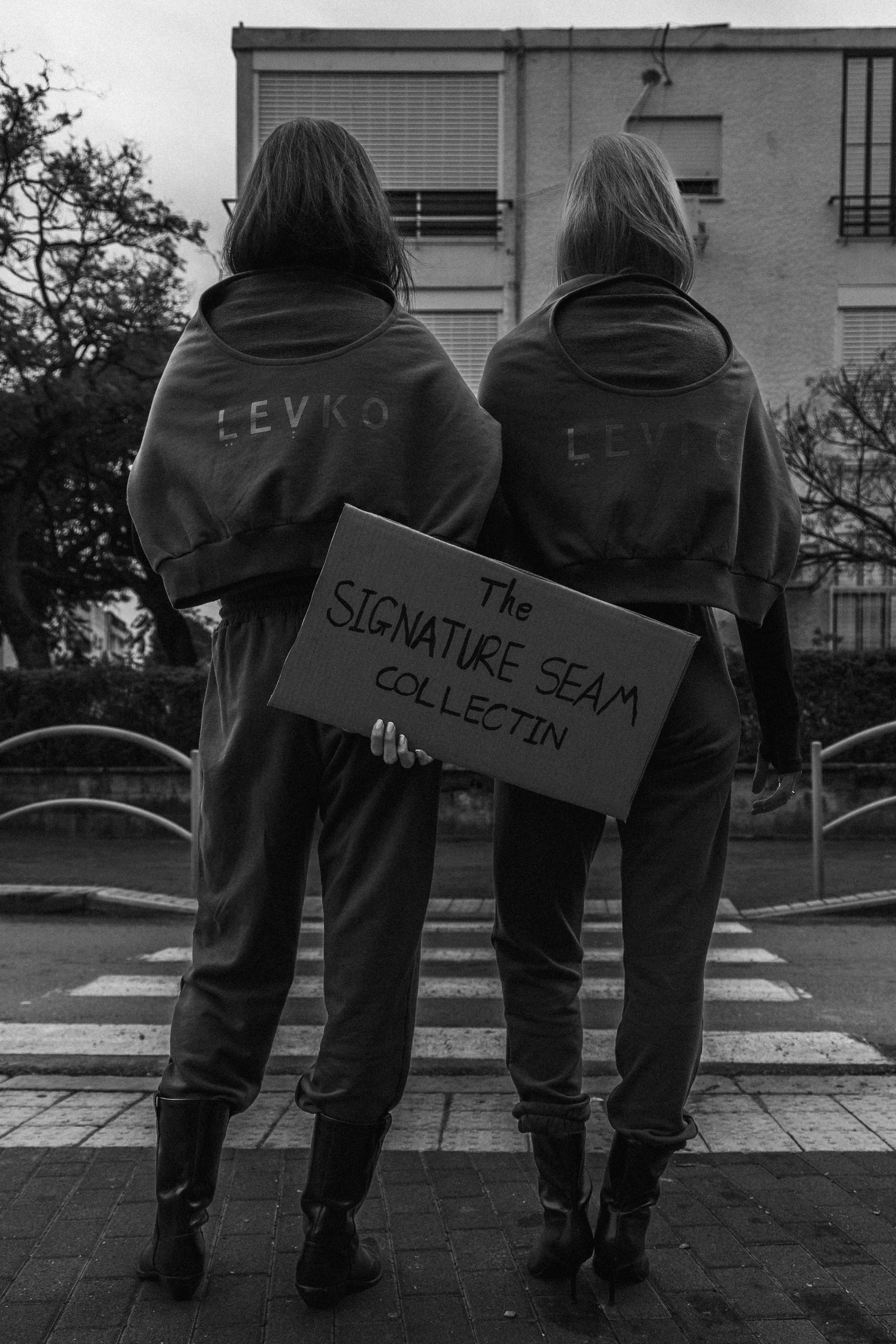 Two women with their backs facing the camera stand outdoors on a crosswalk, wearing matching dark Levi's sweatshirts and boots. One holds a cardboard sign reading 'The Signature Seam Collectin'.'
