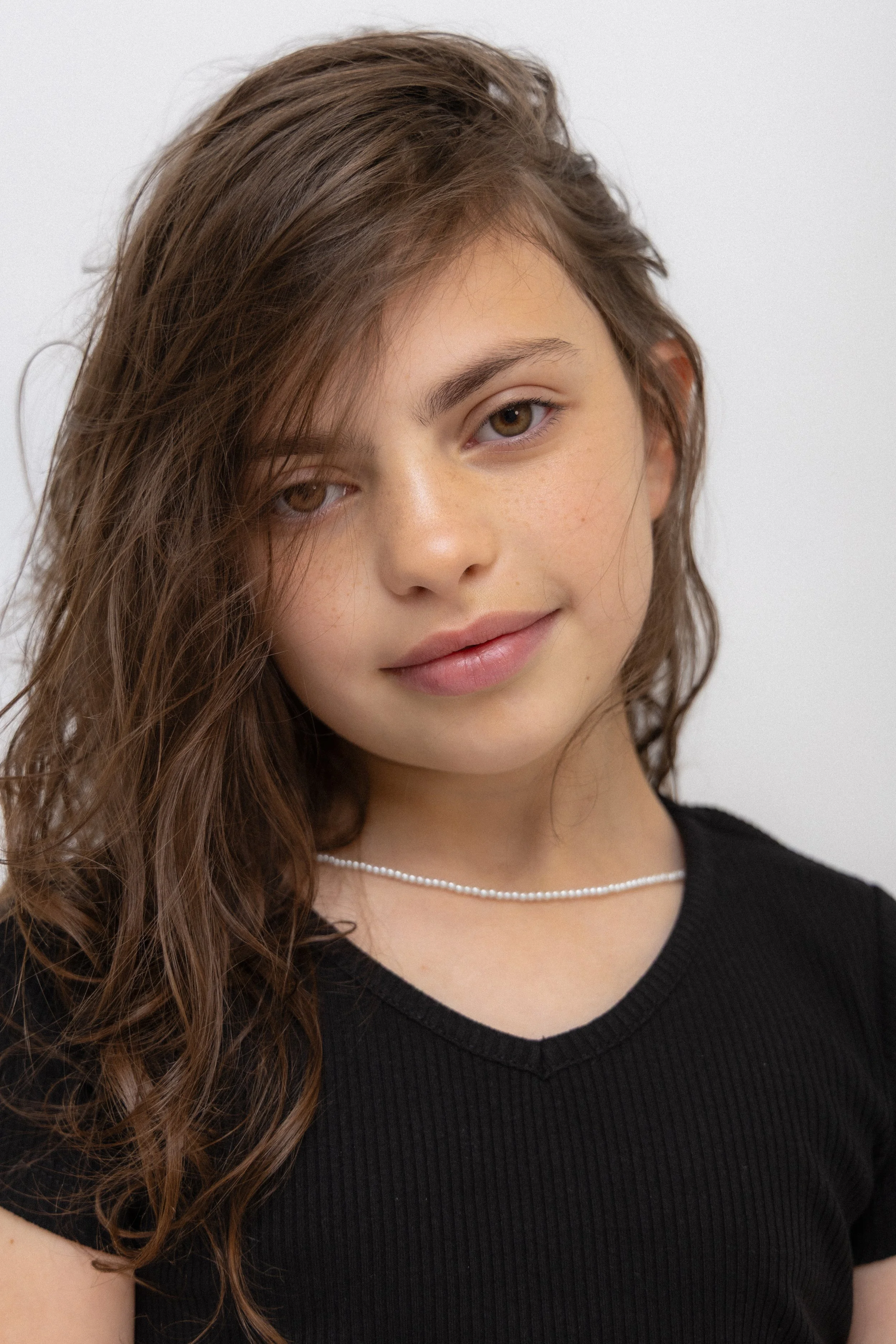 Close-up of a young woman with long, wavy brown hair, wearing a black V-neck top and a delicate pearl necklace, against a plain white background.