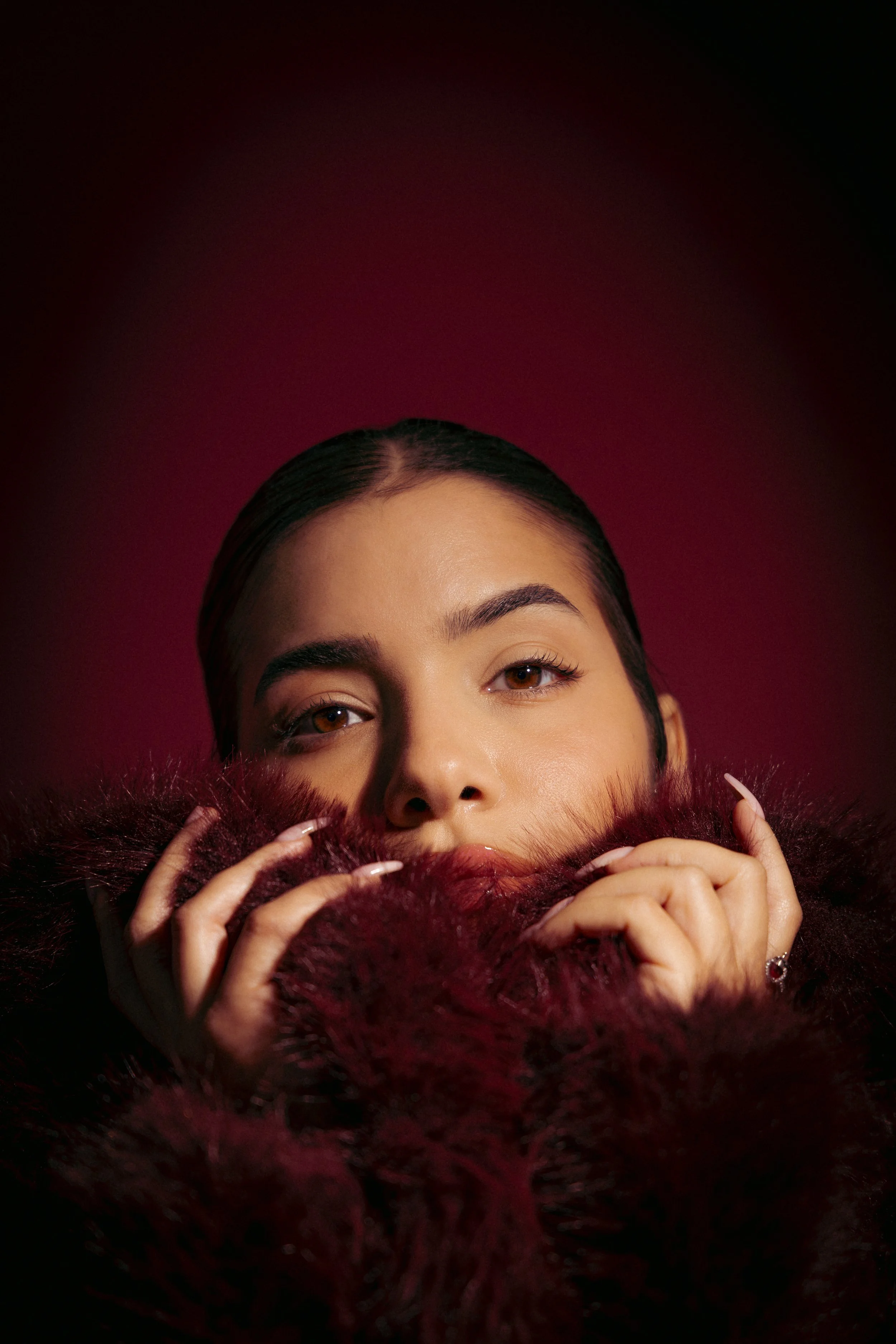 Close-up of a woman with dark hair, brown eyes, and light skin, holding a deep red furry coat around her face, set against a dark pink background.fashion photography. צילומי אופנה, צילום למותגים, דוגמנית. פורטרייט