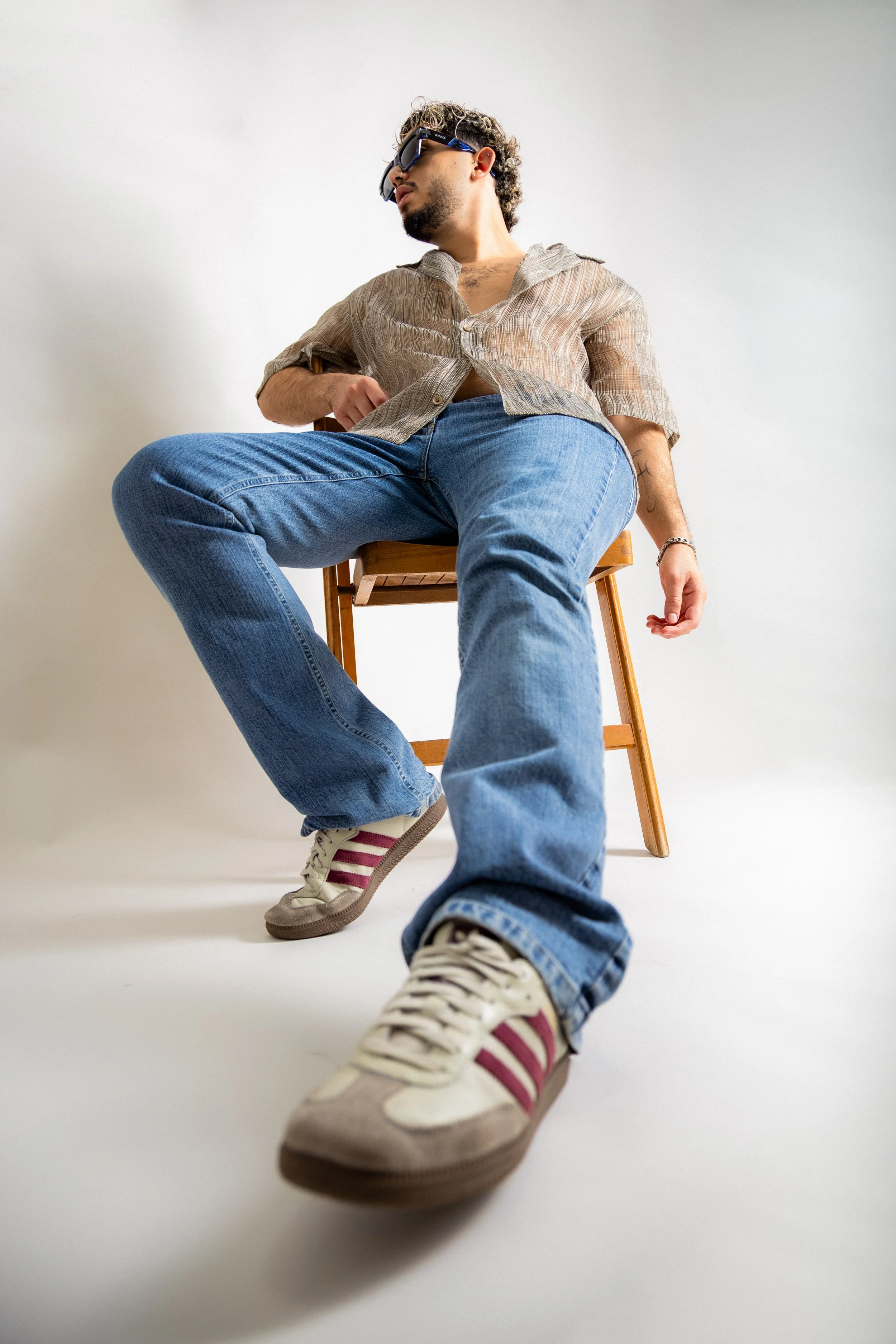 A young man is sitting on a wooden chair against a white background. He has curly hair, sunglasses, and is wearing a partially unbuttoned striped shirt with rolled-up sleeves, blue jeans, and sneakers with pink and white stripes. He is leaning back w