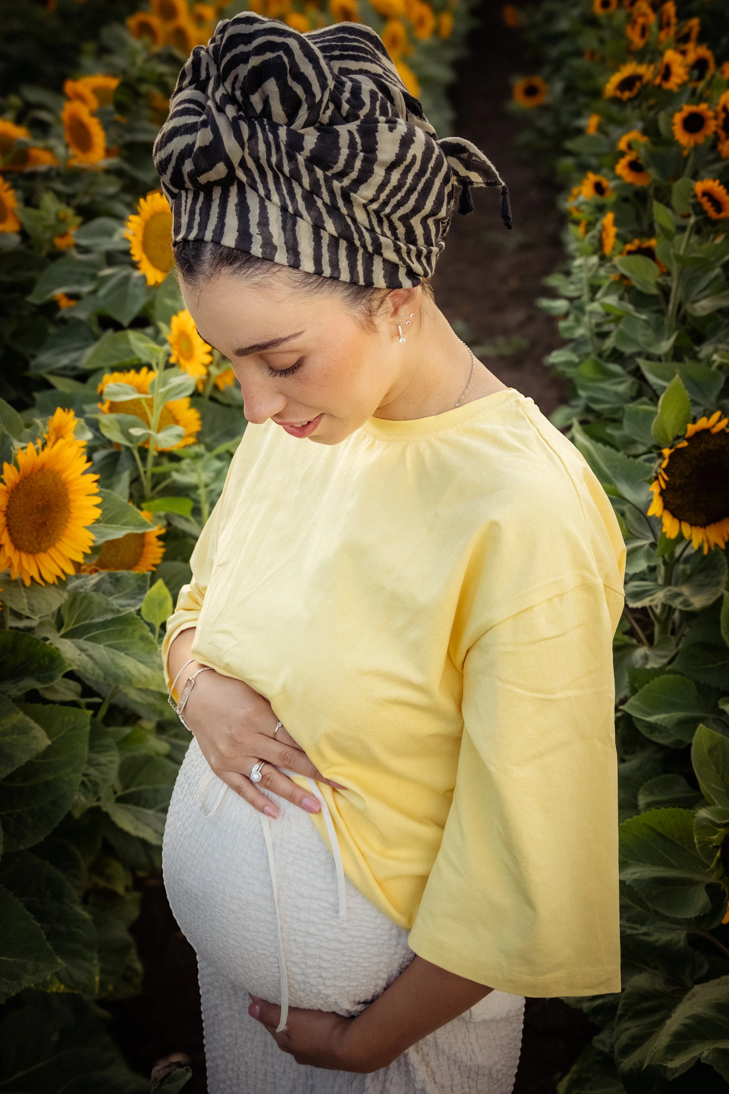 A woman in a zebra-patterned headwrap and yellow shirt holding a baby in white textured clothing among sunflowers.