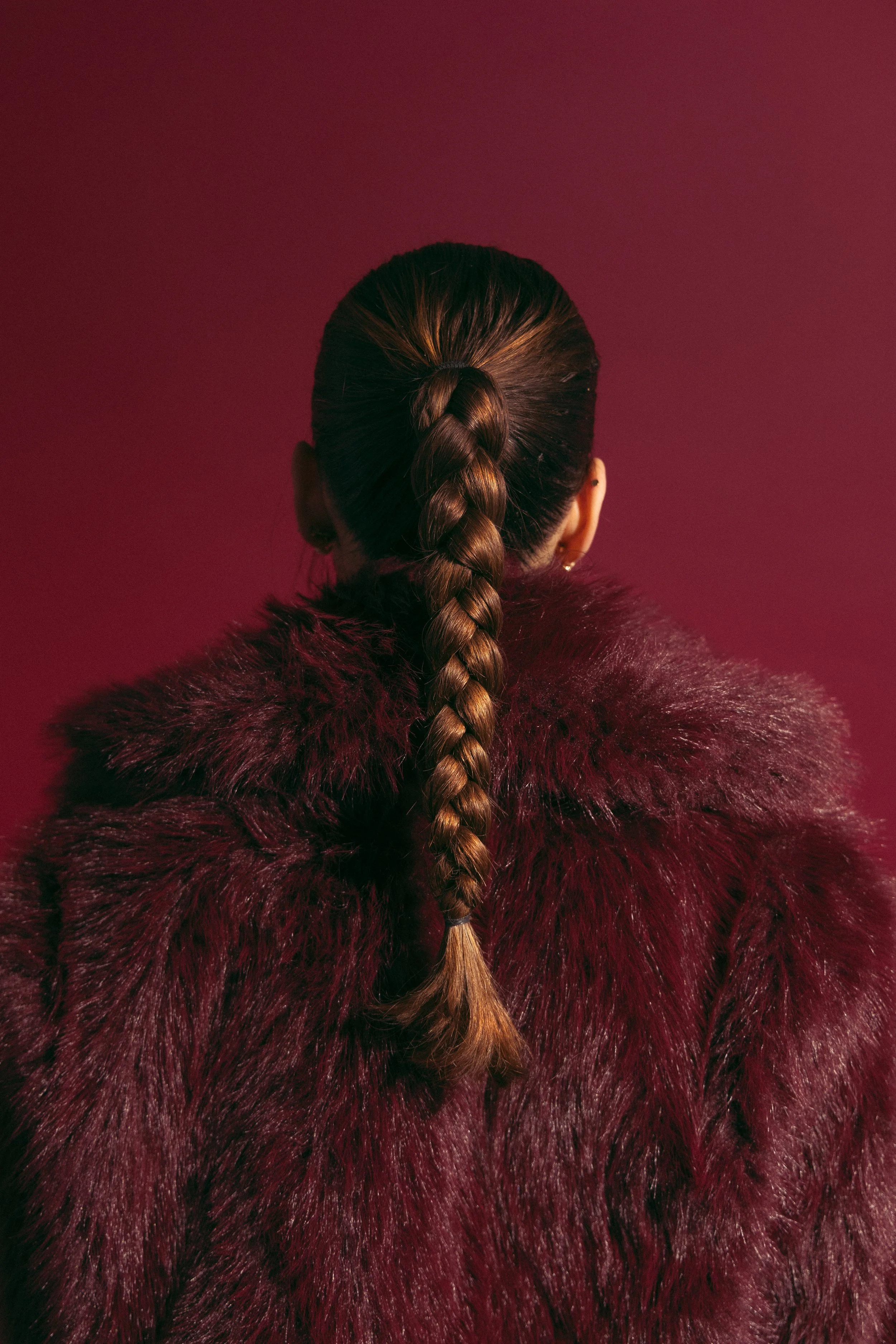 Rear view of a woman with long braided brown hair wearing a burgundy faux fur coat against a matching Burgundy background.