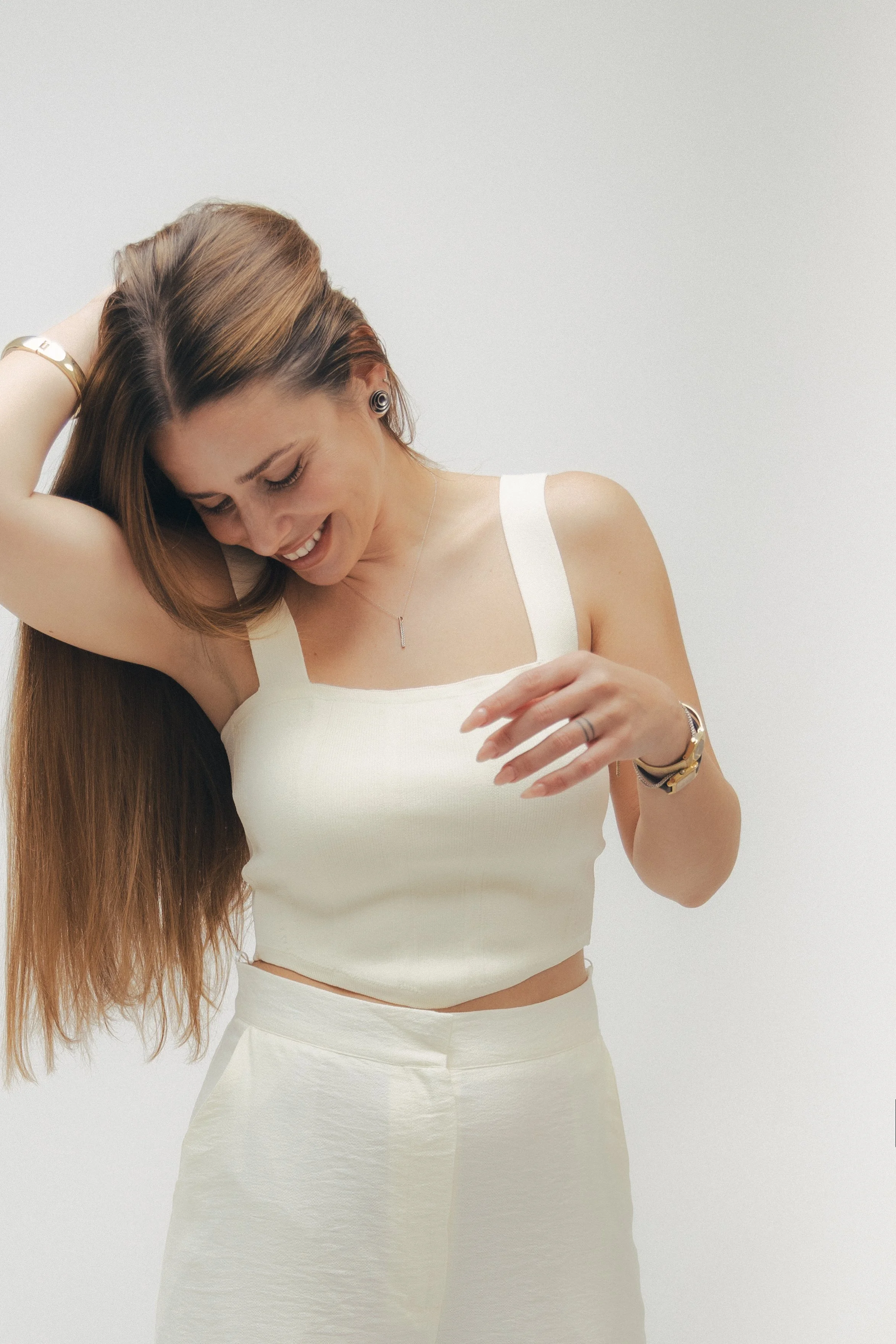 A smiling woman with long brown hair, wearing a white tank top and skirt, adjusting her hair with one hand, standing against a plain white background.