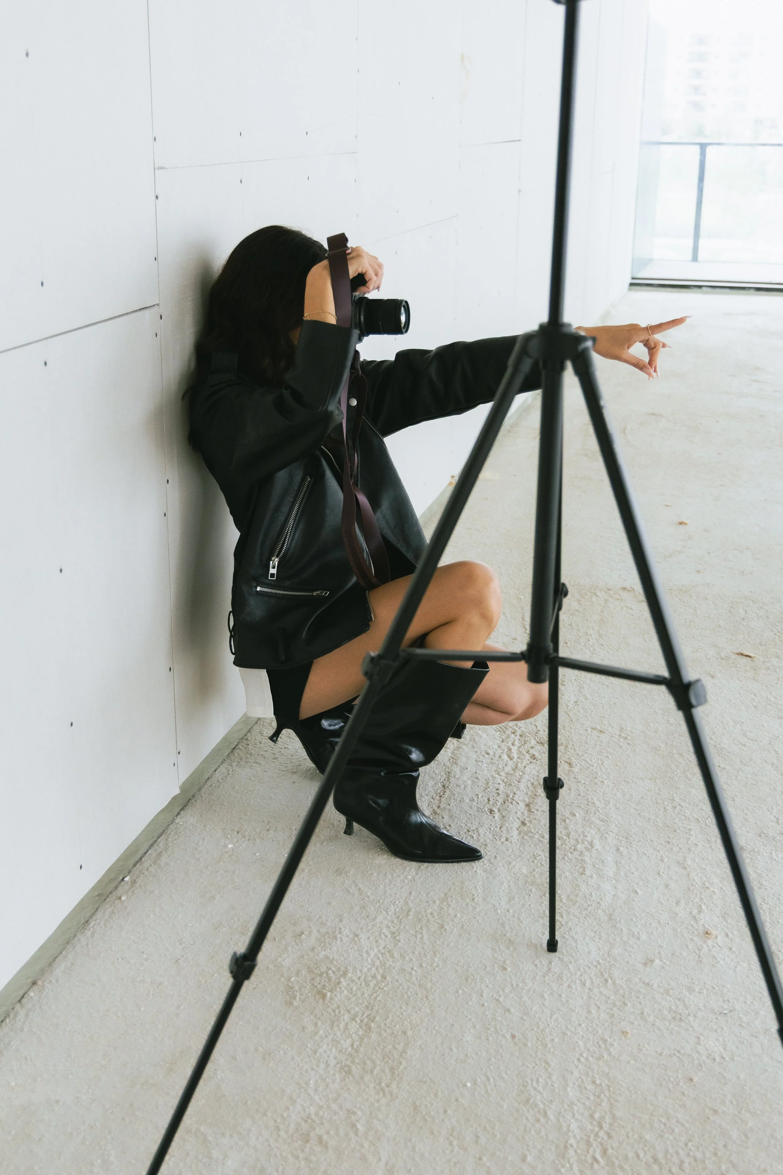 Woman sitting on the floor against a wall, taking a photograph with a camera, and pointing with her right hand.