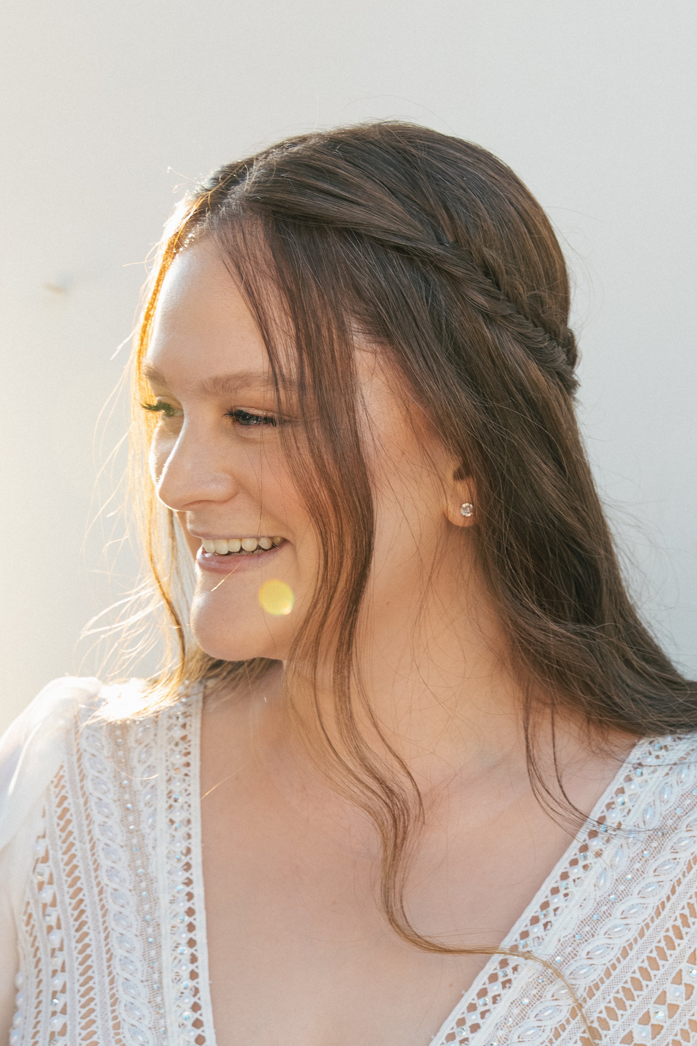 A woman with long brown hair styled in a loose braid, wearing a white lace top and small stud earrings, smiling gently in sunlight.