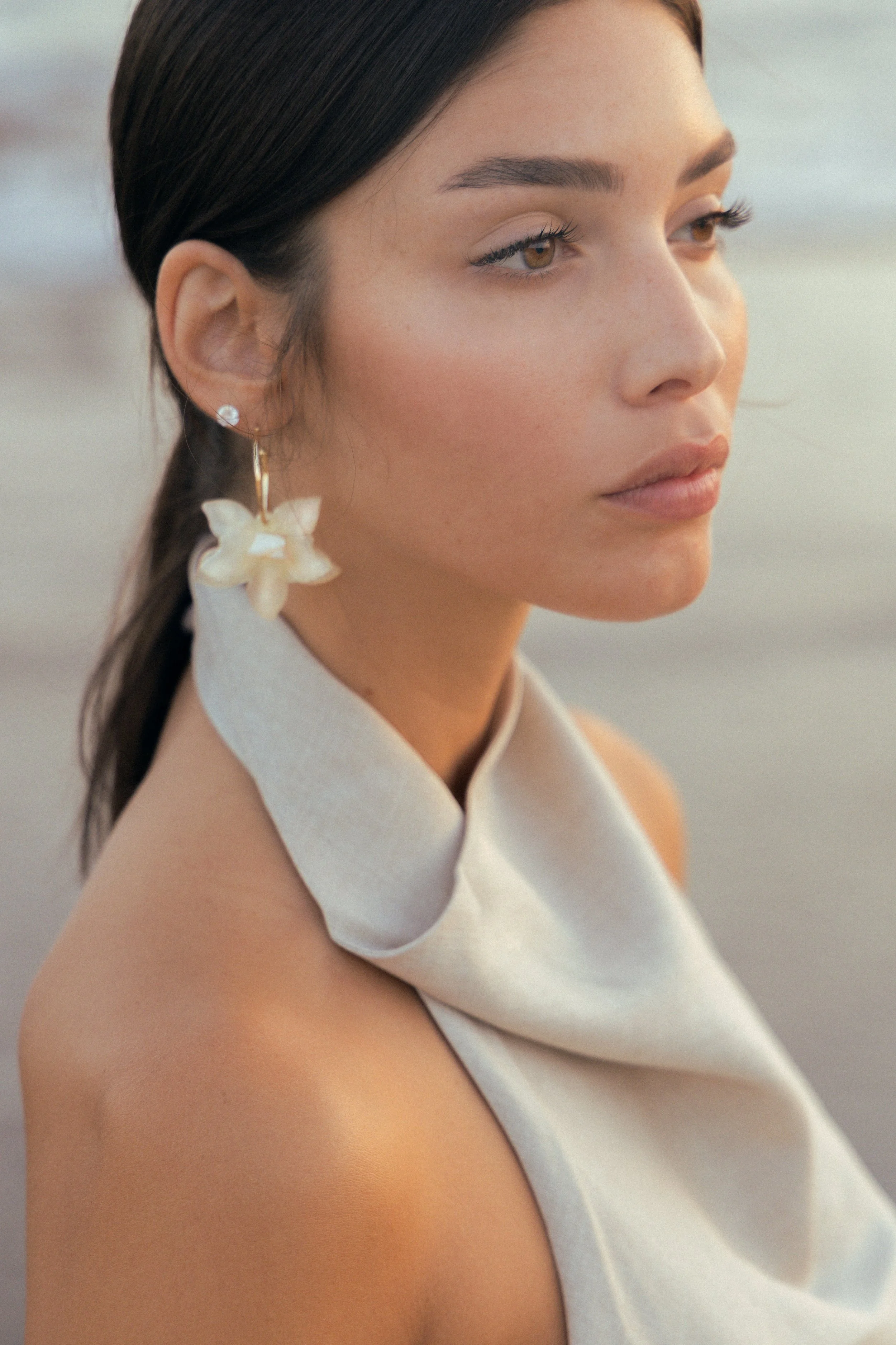Close-up of a woman's face and shoulder, with her hair pulled back, wearing a white dress with a high neckline and a flower-shaped earring. The background is blurred and appears to be outdoors.