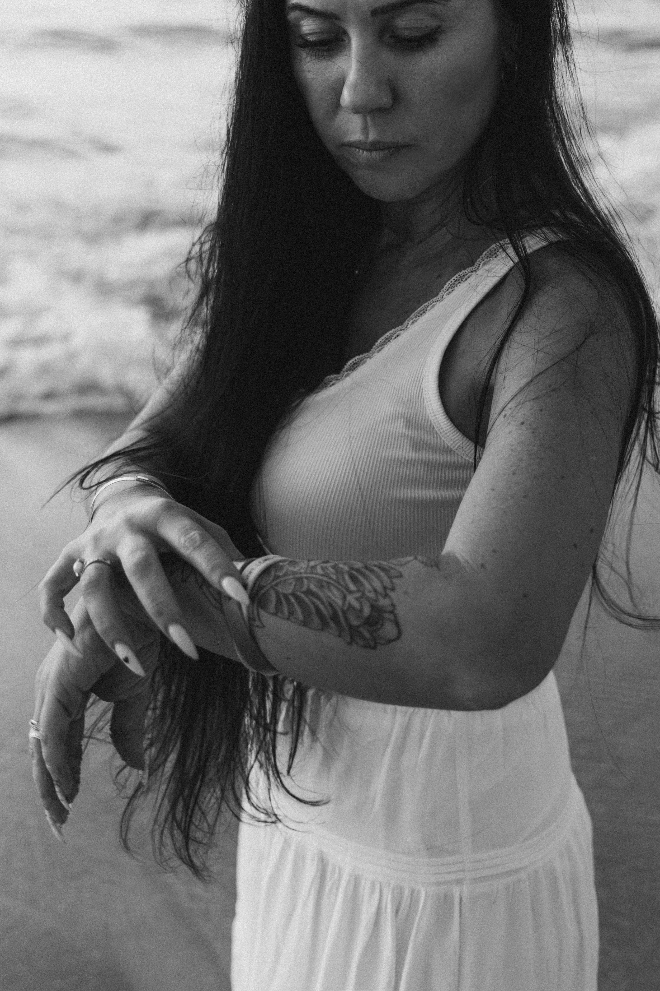 A woman with long dark hair and tattoos on her arms, wearing a white tank top with lace trim and a matching white skirt, looking down while standing outdoors.