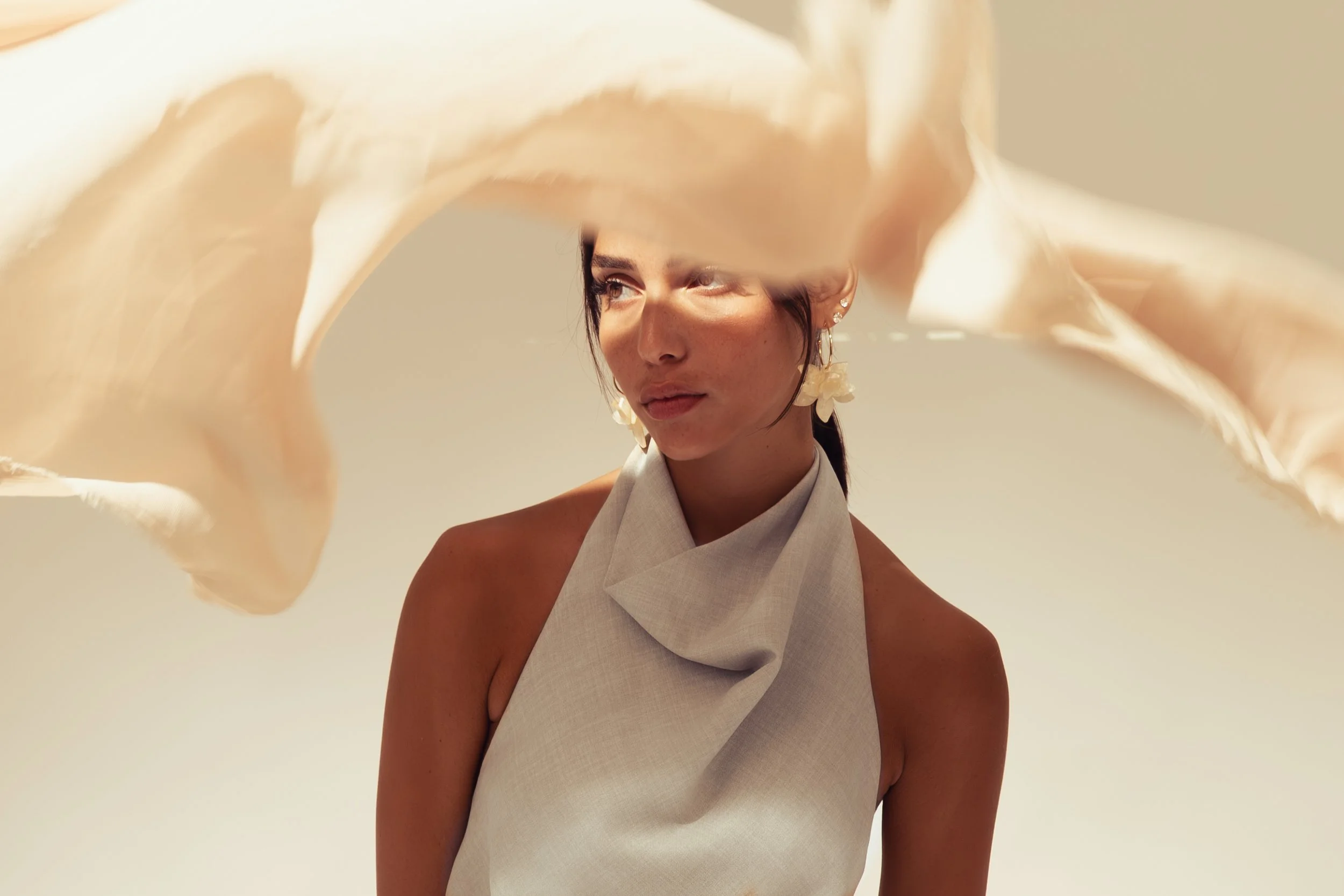 A woman with dark hair, wearing a beige halter top and large flower earrings, is holding a flowing, light-colored fabric overhead, partially obscuring her face and creating a dynamic, artistic composition against a neutral background.