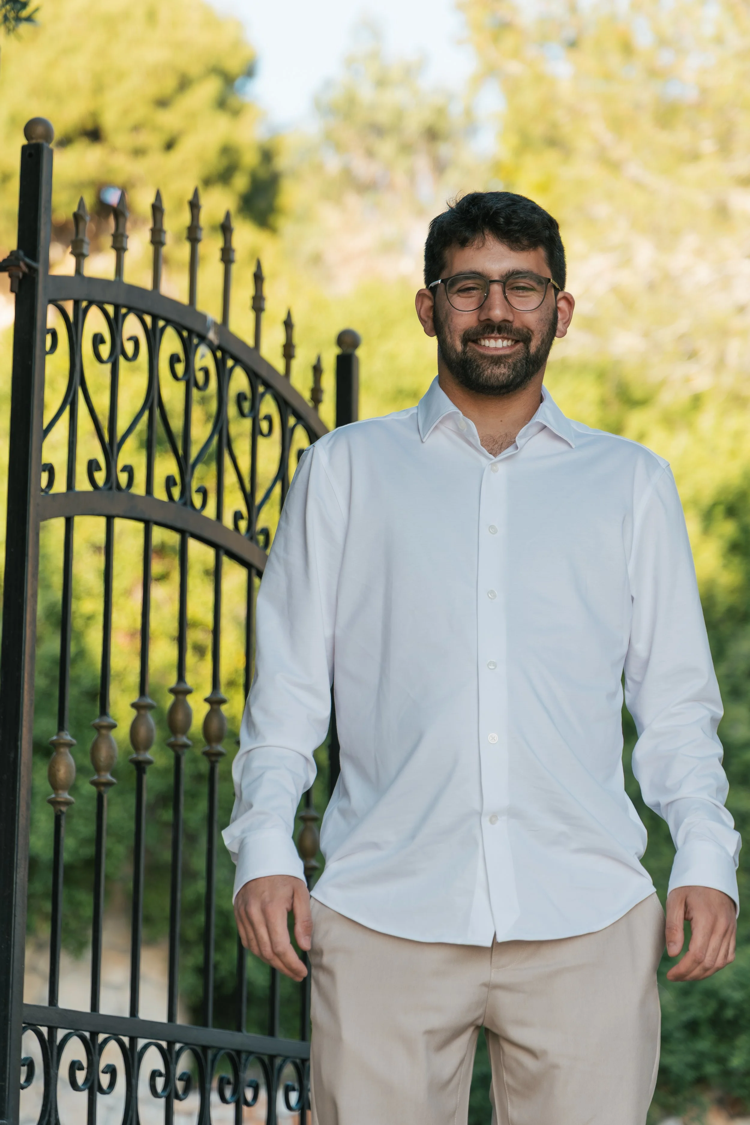 A man with dark hair, glasses, and a beard standing outdoors near a black wrought iron gate, smiling, wearing a white dress shirt and beige pants, with blurred trees in the background.