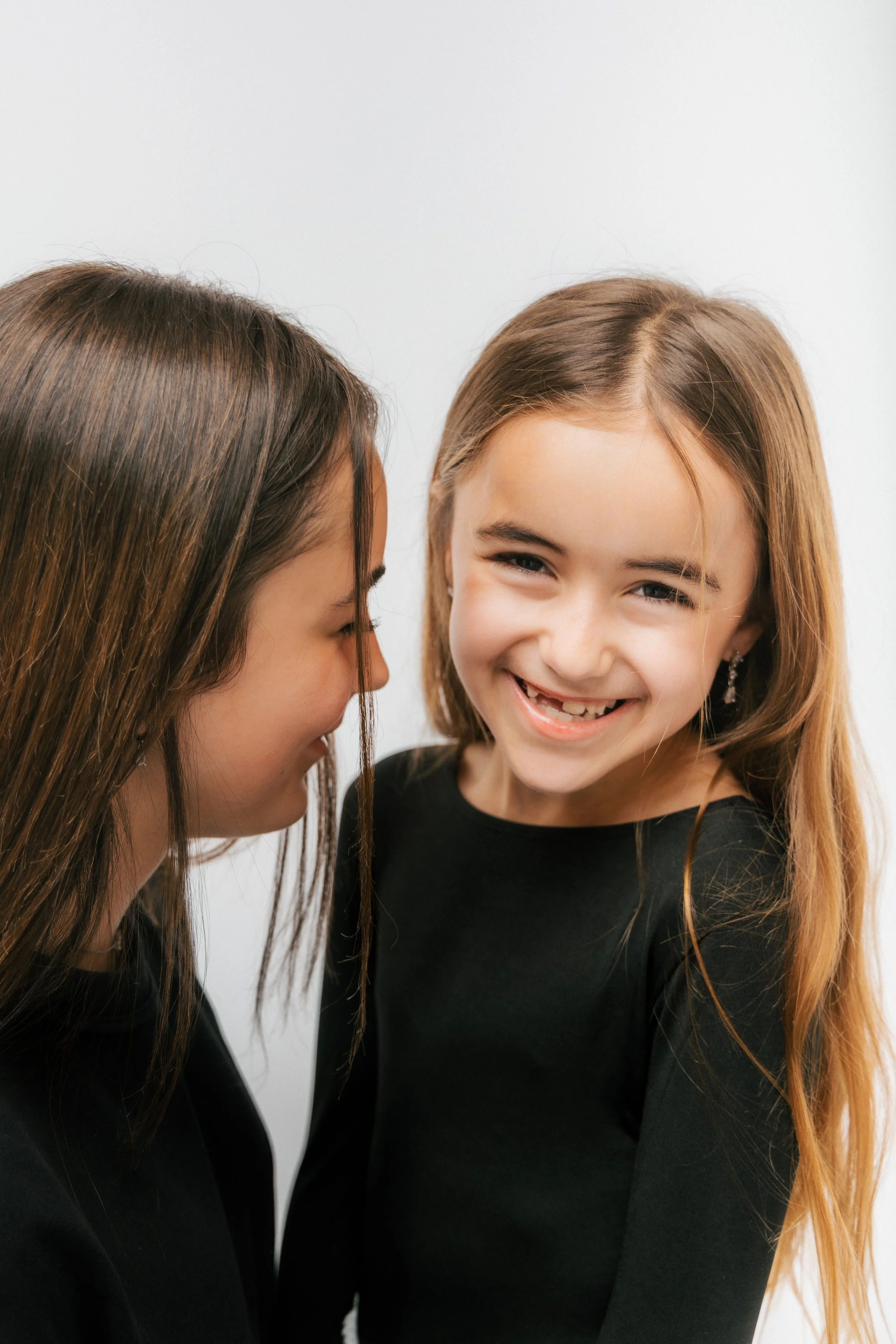 Two young girls with long hair, wearing black shirts, sharing a private joke or whispering to each other, smiling with joy, against a plain white background.