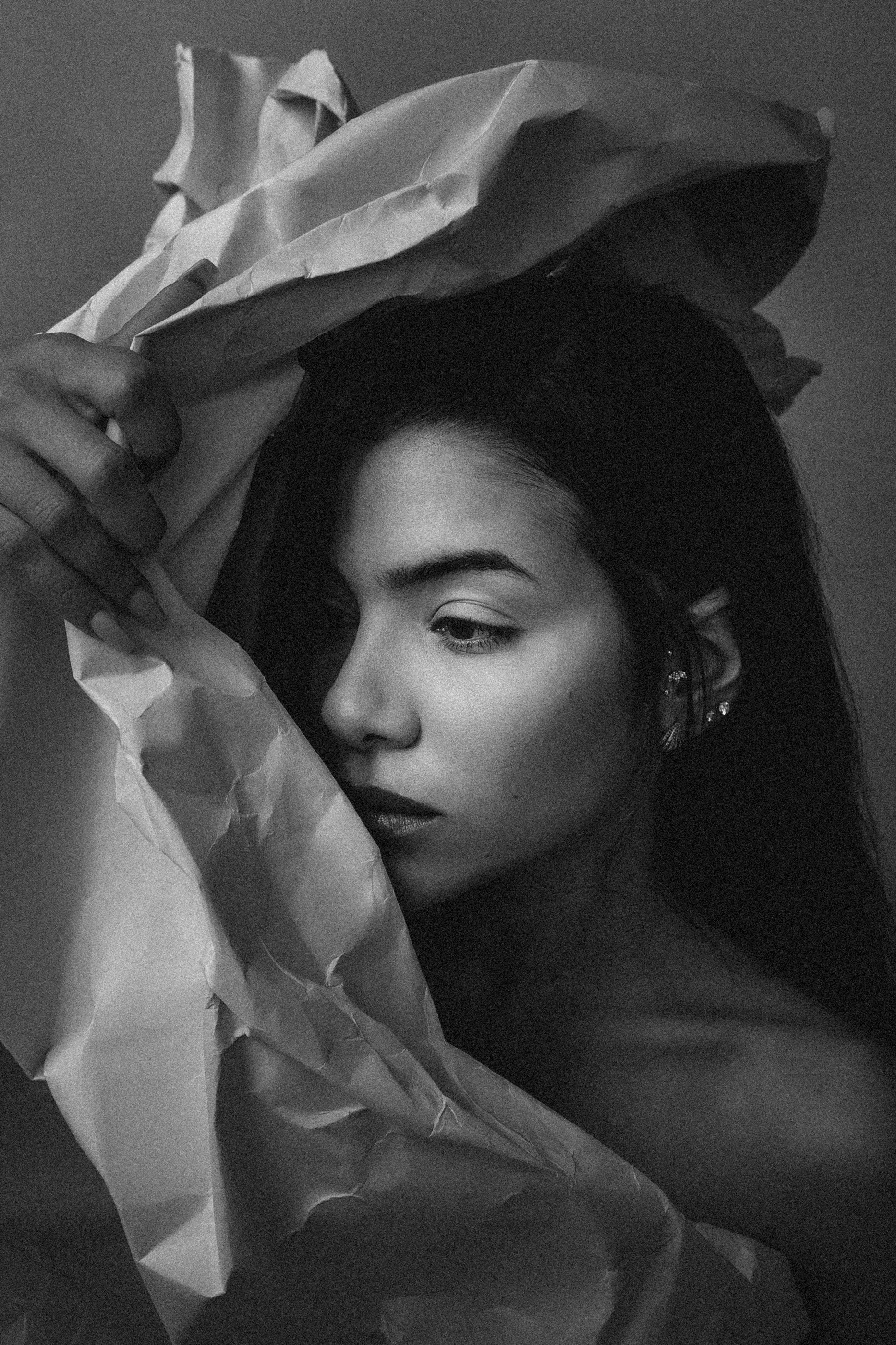 A black and white photo of a woman with dark hair and earrings holding crumpled paper near her face, partially obscuring her features.