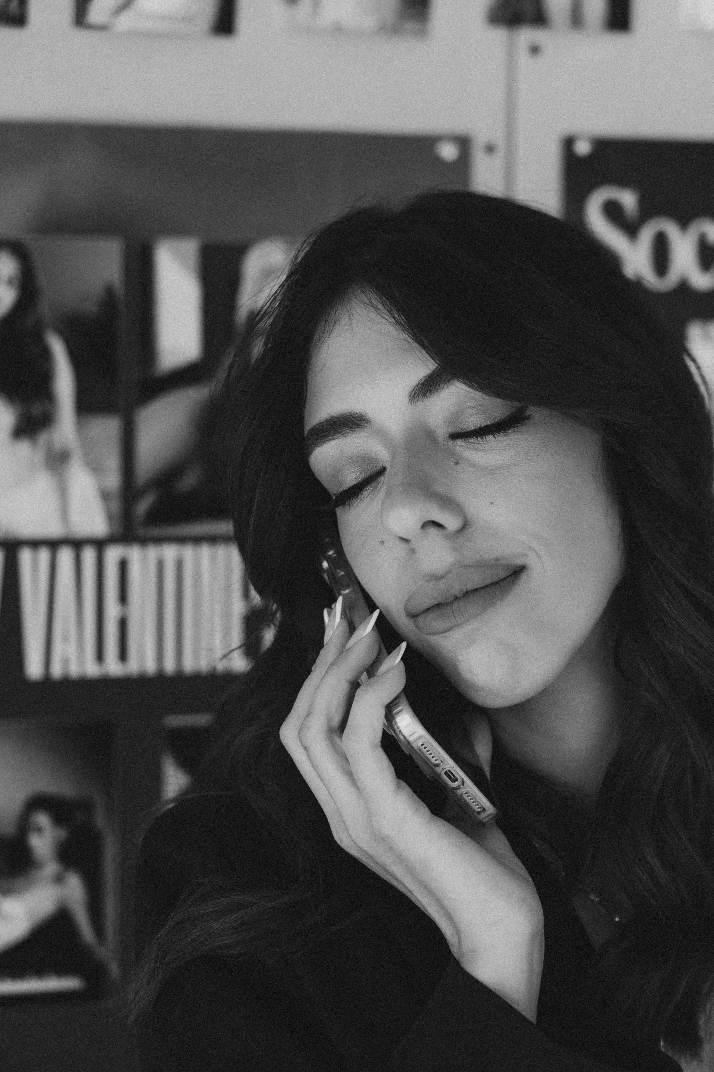 A woman with dark hair and light makeup smiling with eyes closed, on the phone, in a school or office environment with bookshelves and posters in the background.