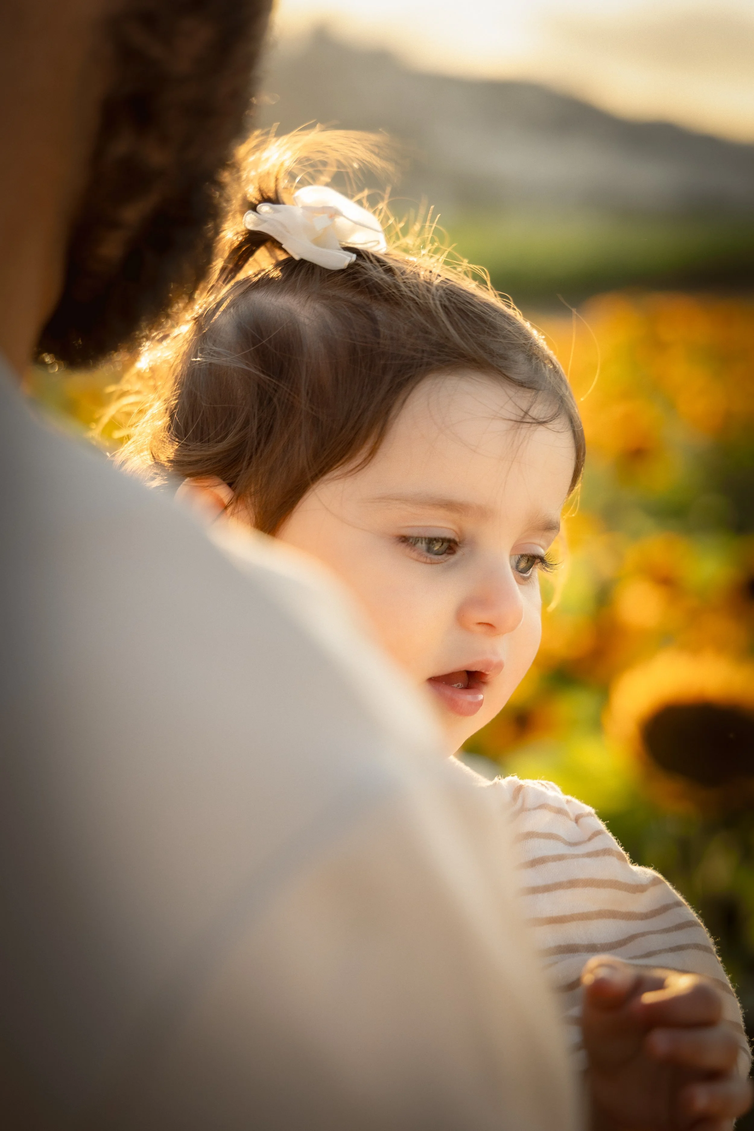 A young girl with brown hair and green eyes is outdoors in sunlight, wearing a white and beige striped shirt, with a white bow in her hair, looking down thoughtfully.