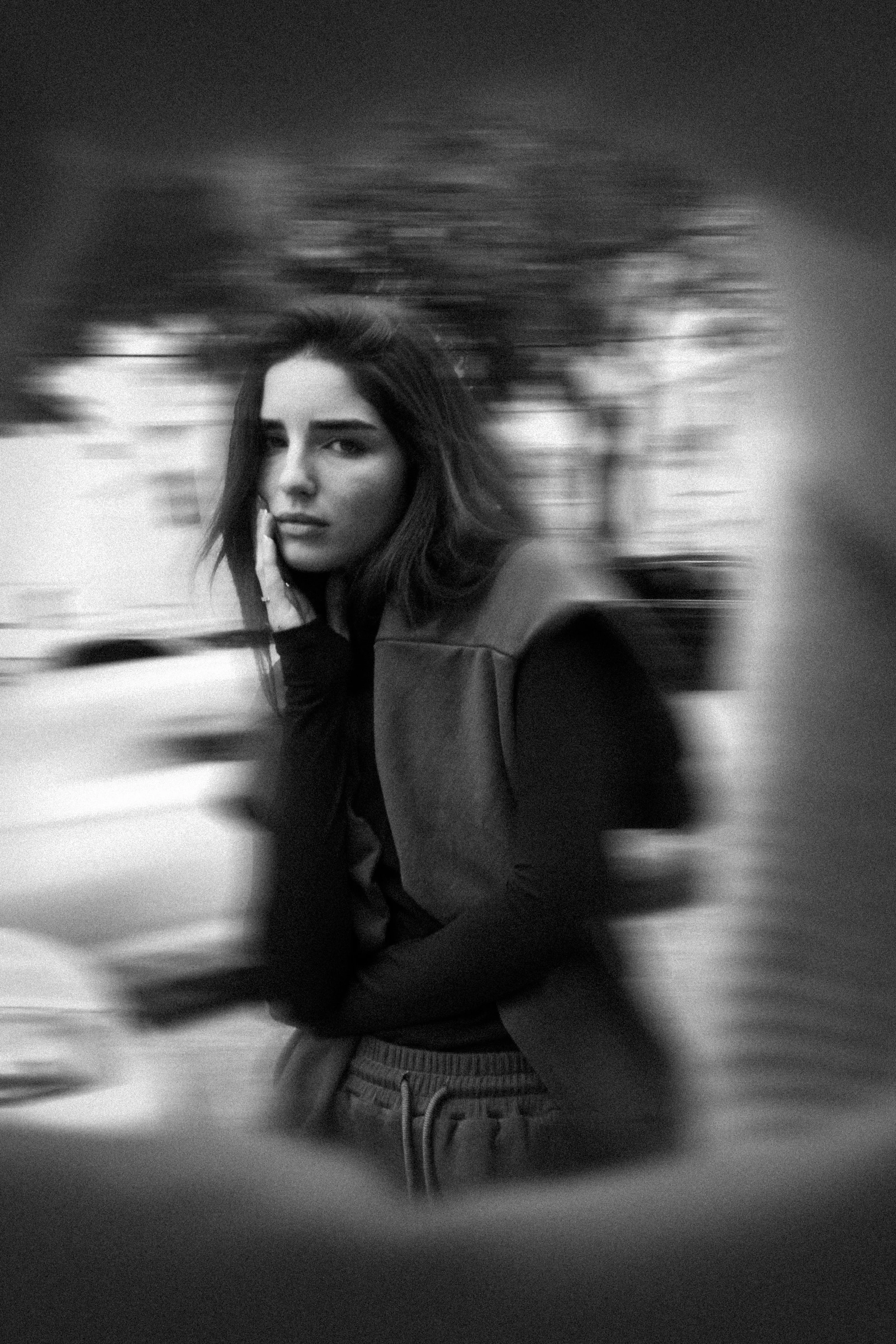 Black and white photo of a young woman with medium-length hair, wearing a dark jacket and striped pants, standing outdoors with blurred trees and sky in the background, looking into the camera with a serious expression.