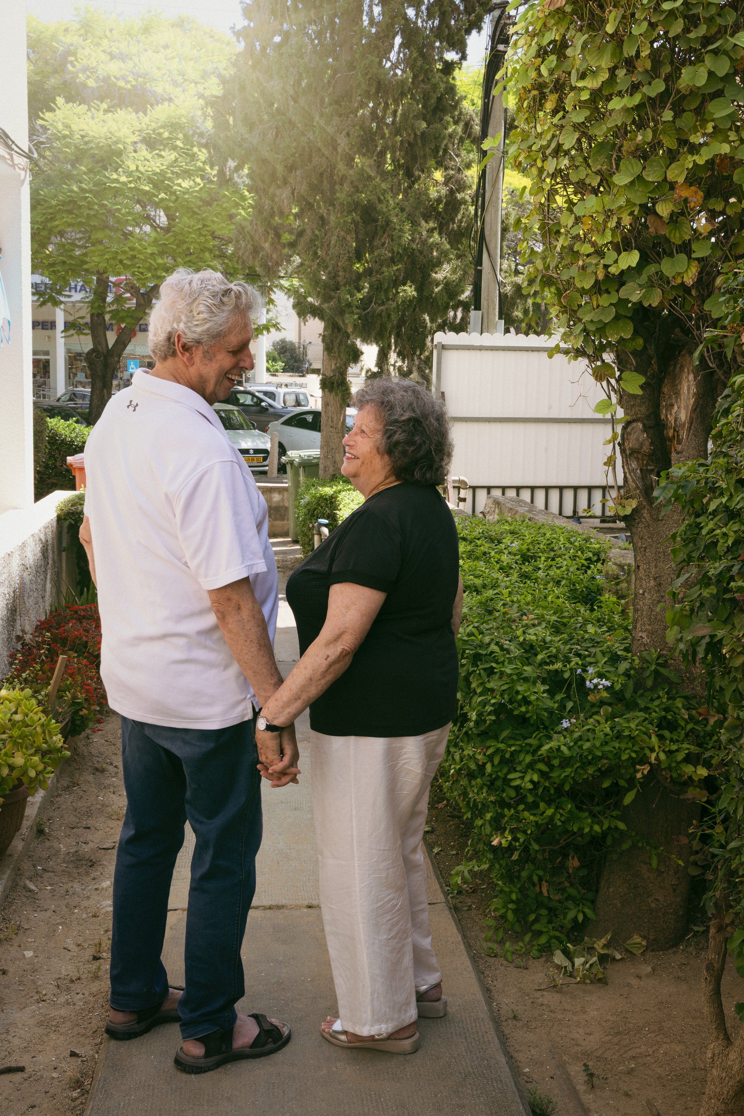 An elderly man and woman holding hands and smiling at each other on a sidewalk surrounded by greenery and trees in a sunny outdoor setting.