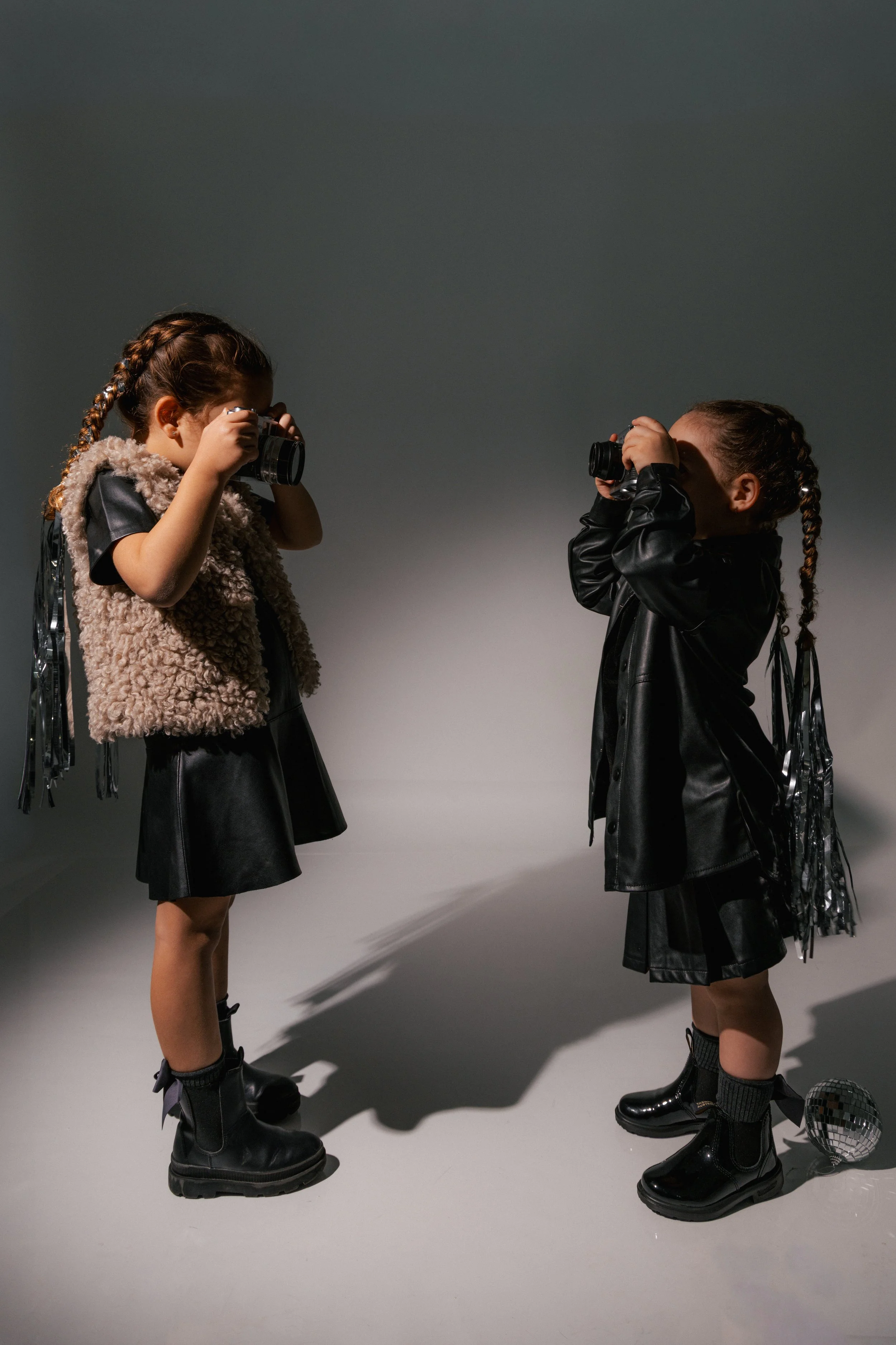 Two young girls standing and facing each other, taking photos with cameras. They are dressed in black outfits with chunky black boots, and have braided hair. One girl is wearing a fluffy vest and the other a shiny black jacket. There are two disco ba