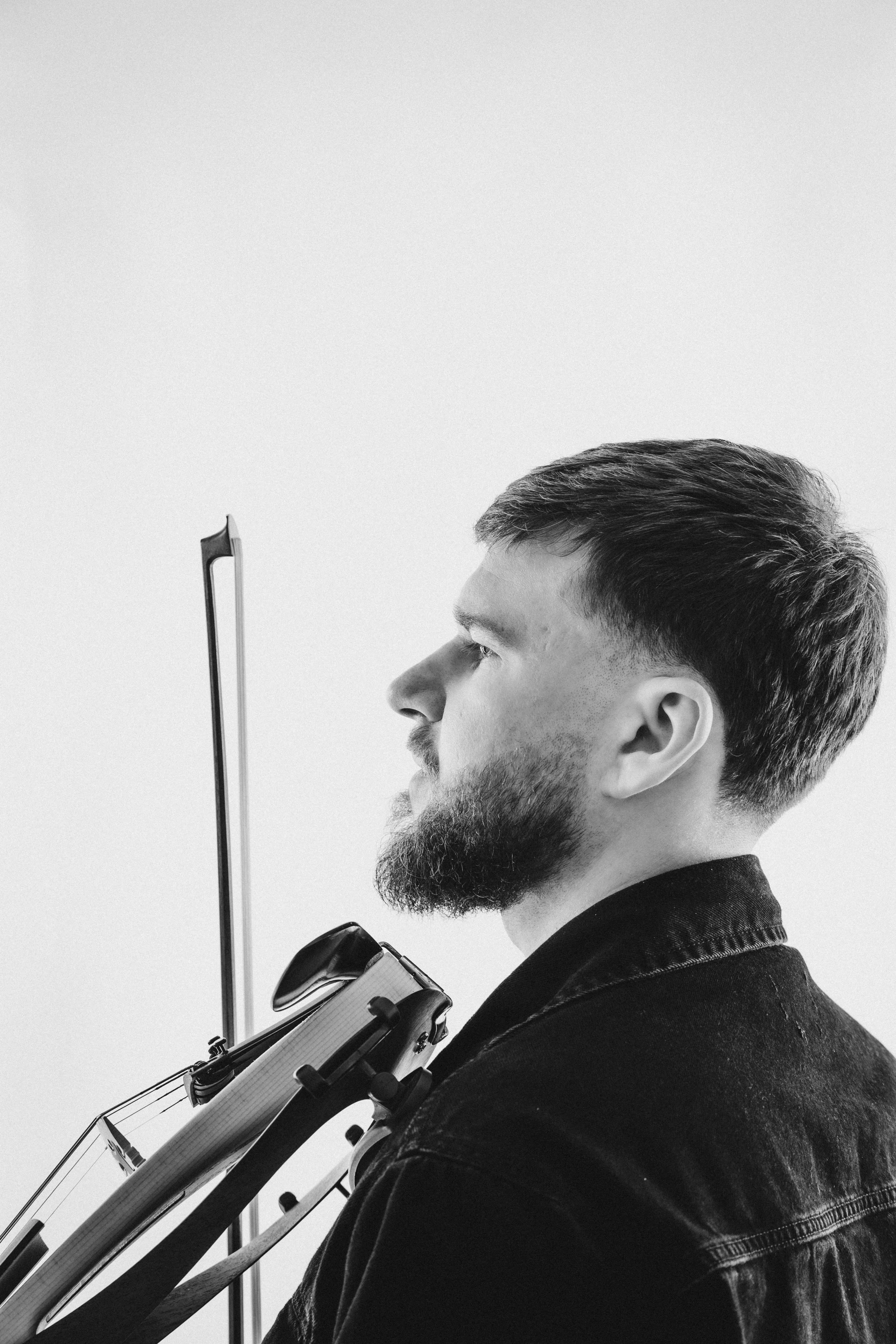 A black and white photo of a man with a beard and short hair holding a violin and bow, facing to the left, against a plain background.