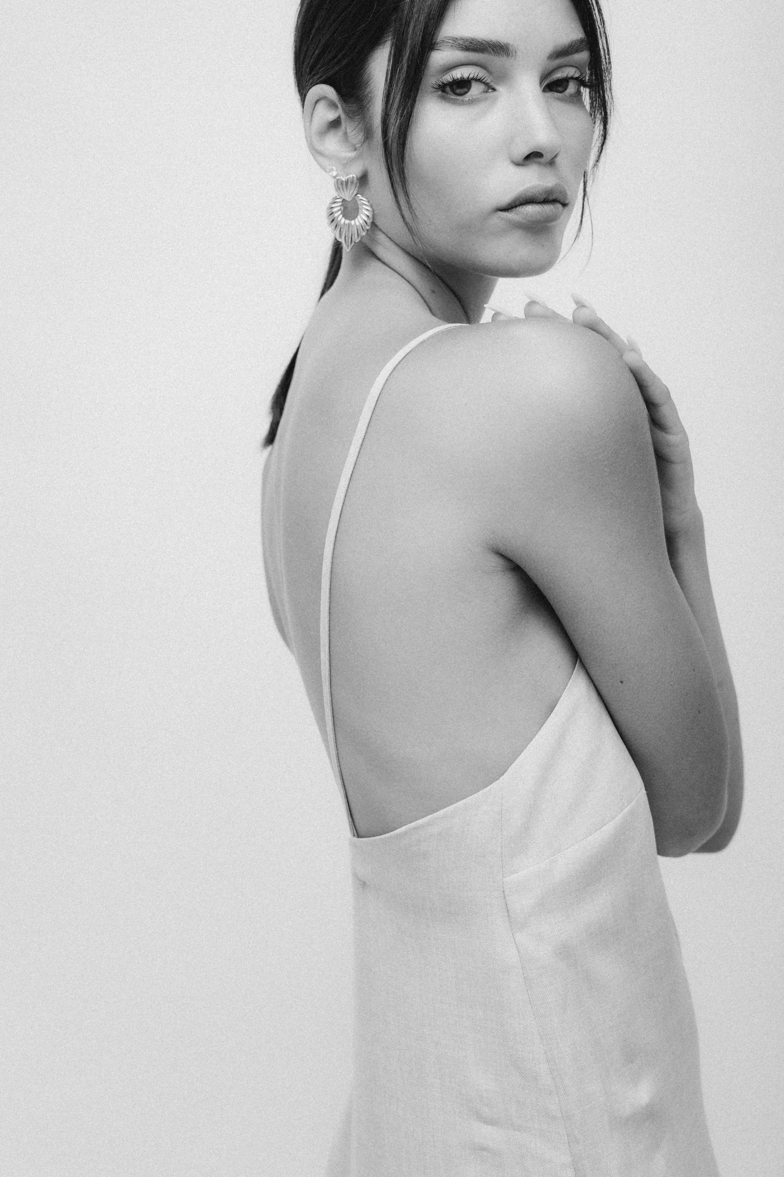 Black and white photo of a woman with dark hair, wearing large earrings and a sleeveless dress, looking over her shoulder with her hand on her shoulder.