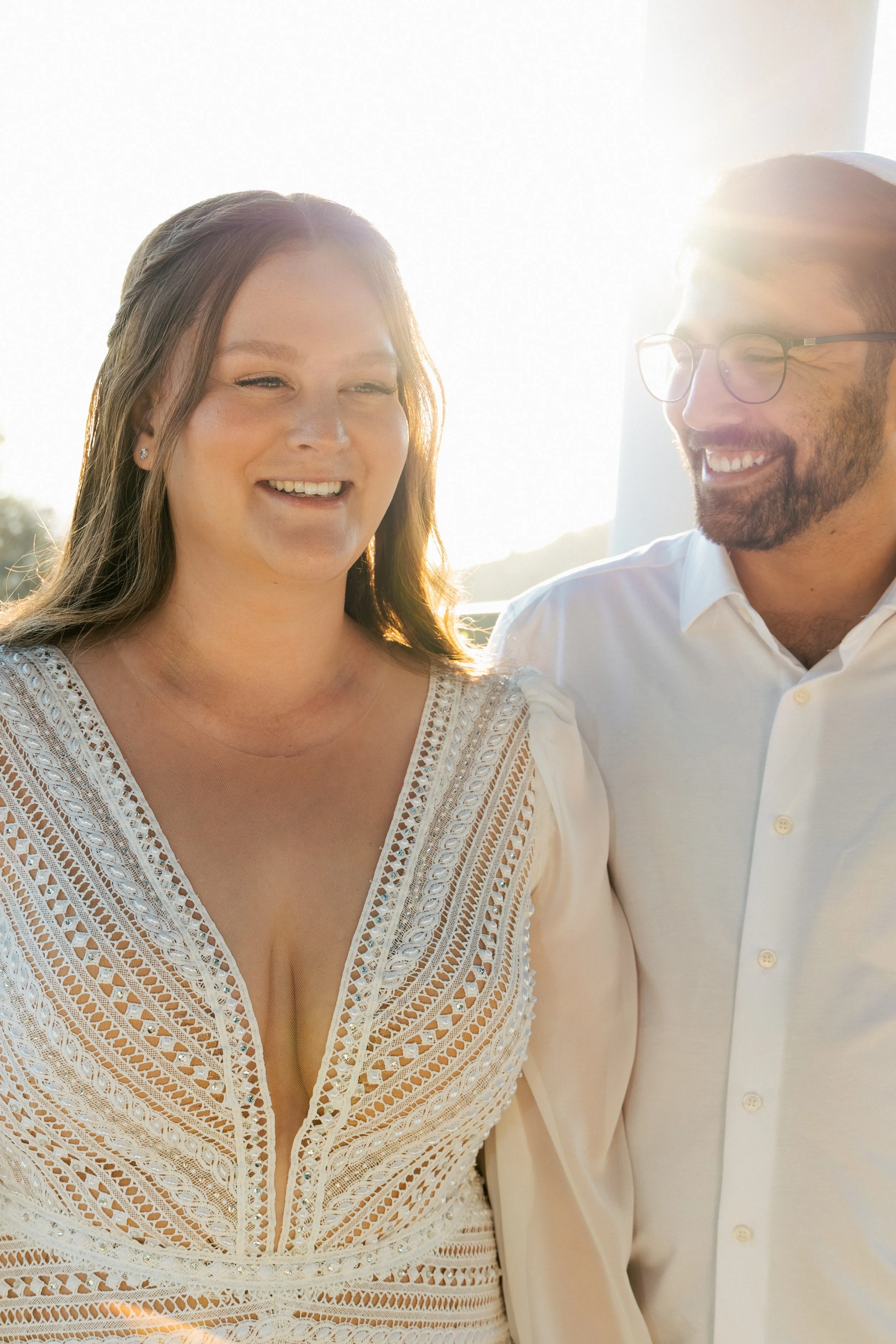 Two people smiling and looking at each other during a sunny outdoor setting, with a woman on the left wearing a white embroidered top and a man on the right wearing glasses and a white shirt.