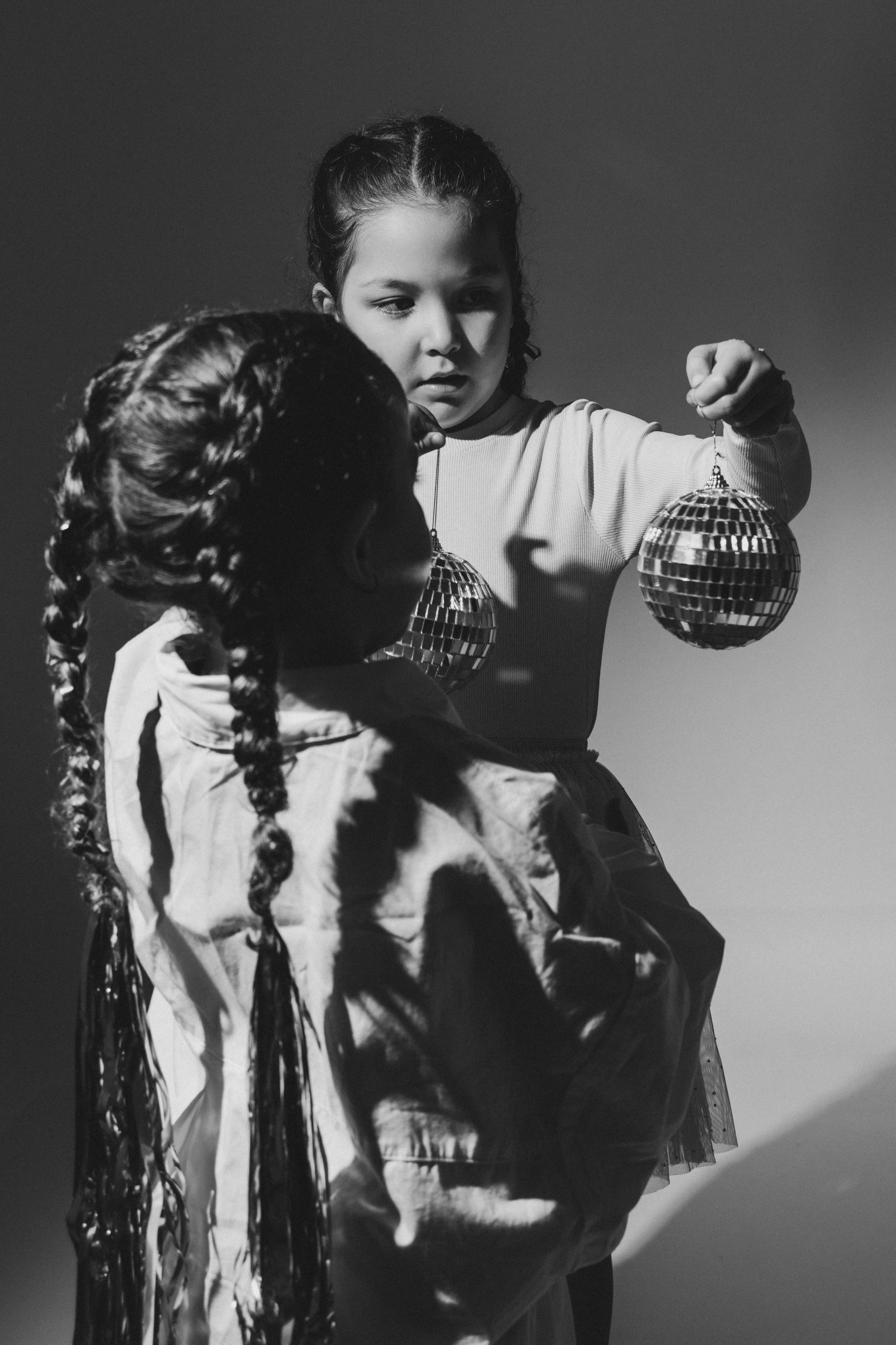 Two young girls with braids holding jewelry or ornaments, with one girl looking at the ornaments and the other girl facing away, in black and white.