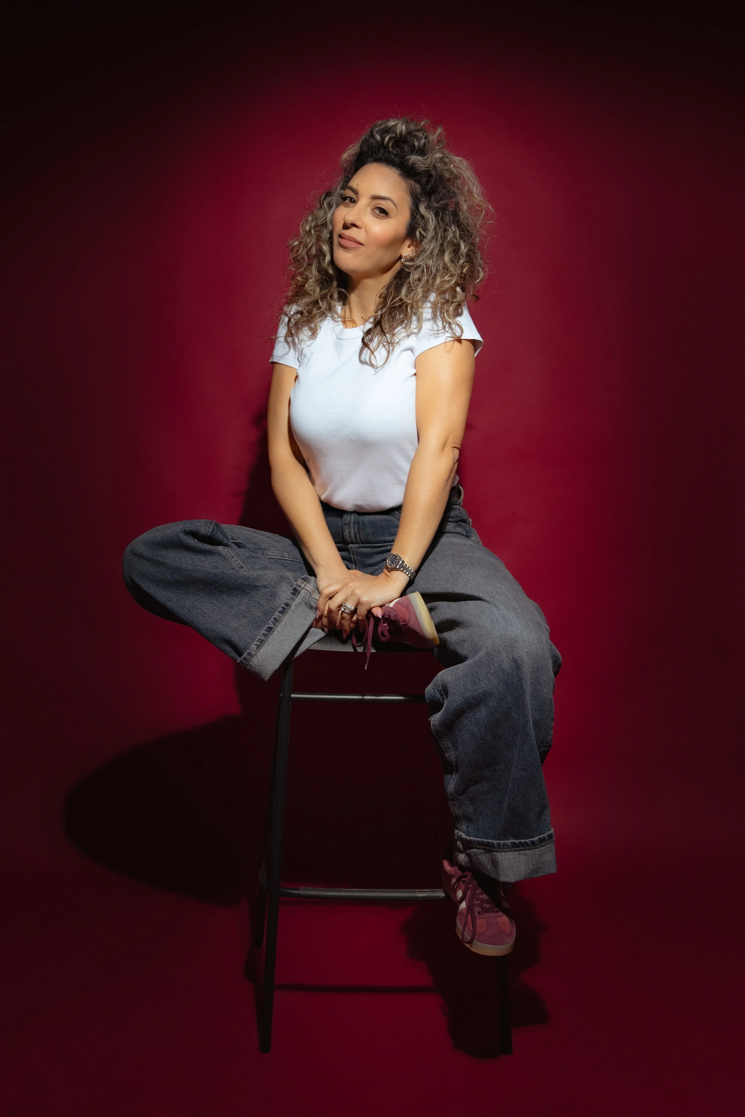 A woman with curly hair sitting on a high stool, wearing a white T-shirt, baggy jeans, maroon sneakers, and a watch, against a dark red background.