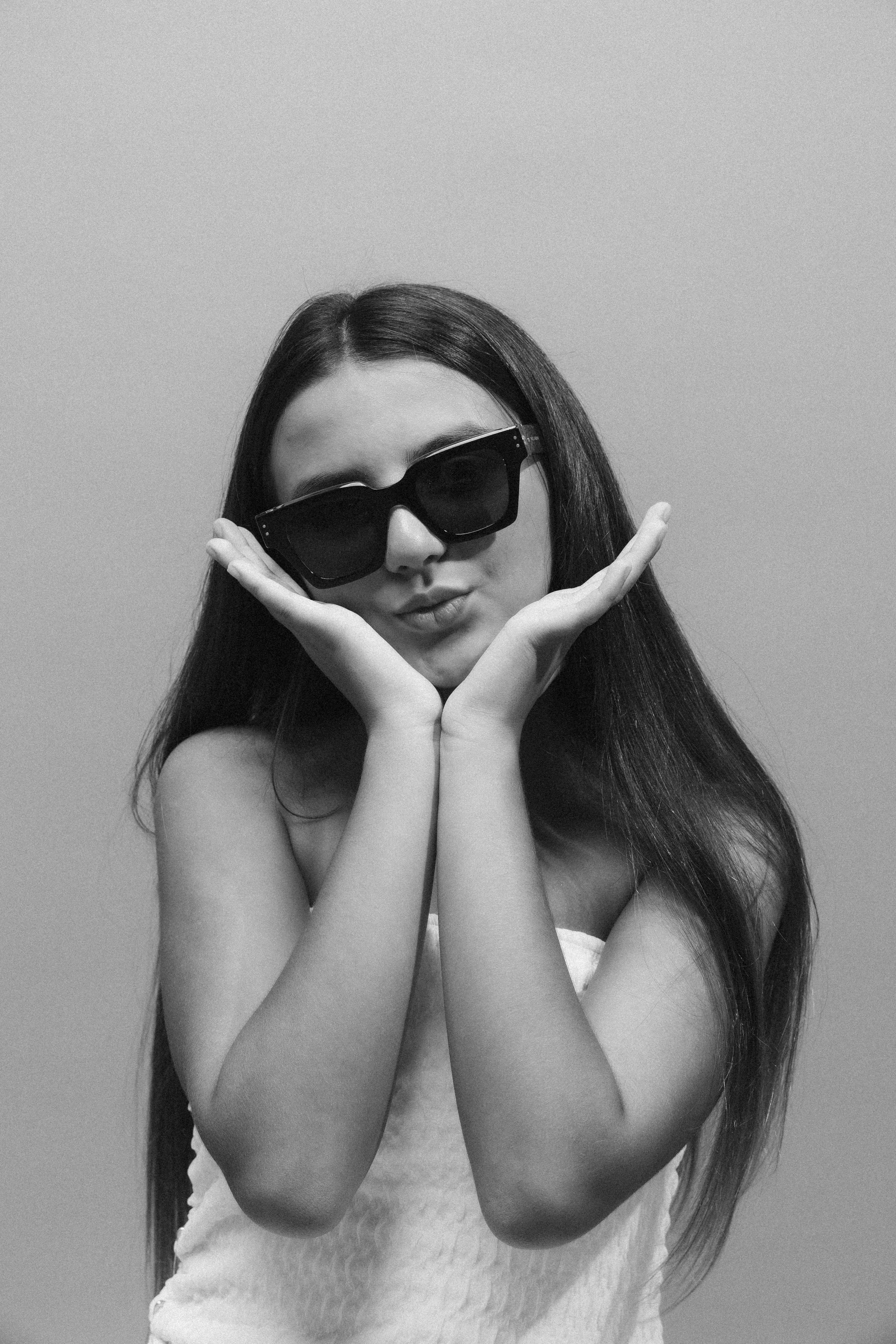 A woman with long dark hair wearing large sunglasses and a strapless top, posing with her palms under her chin and lips pursed against a plain background.
