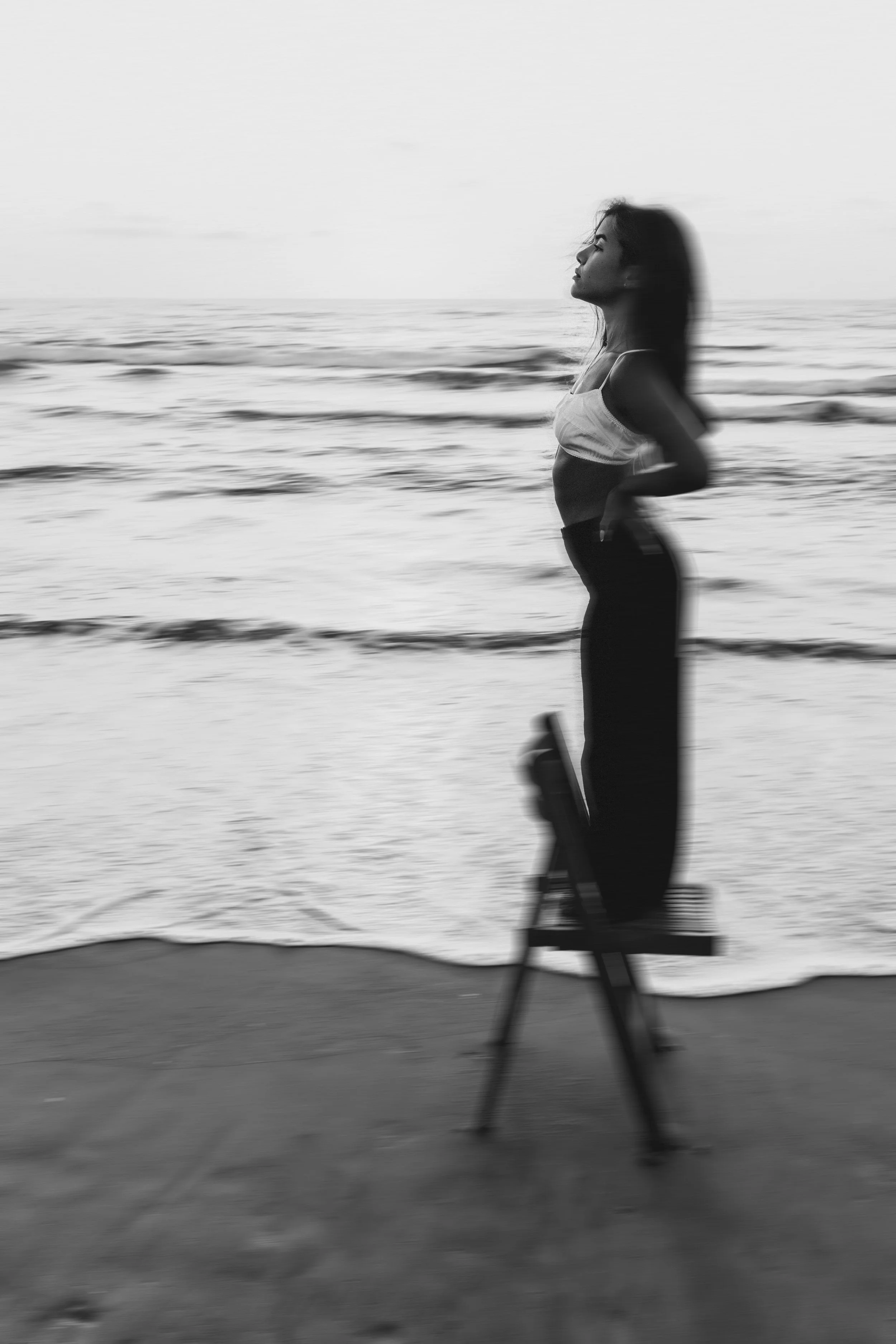 A black-and-white photo of a woman with long hair in profile, sitting in a chair facing the ocean, with her eyes closed and a calm expression, near the beach with waves in the background. artistic edit. model. outdoor photoshoot. צילומים בים