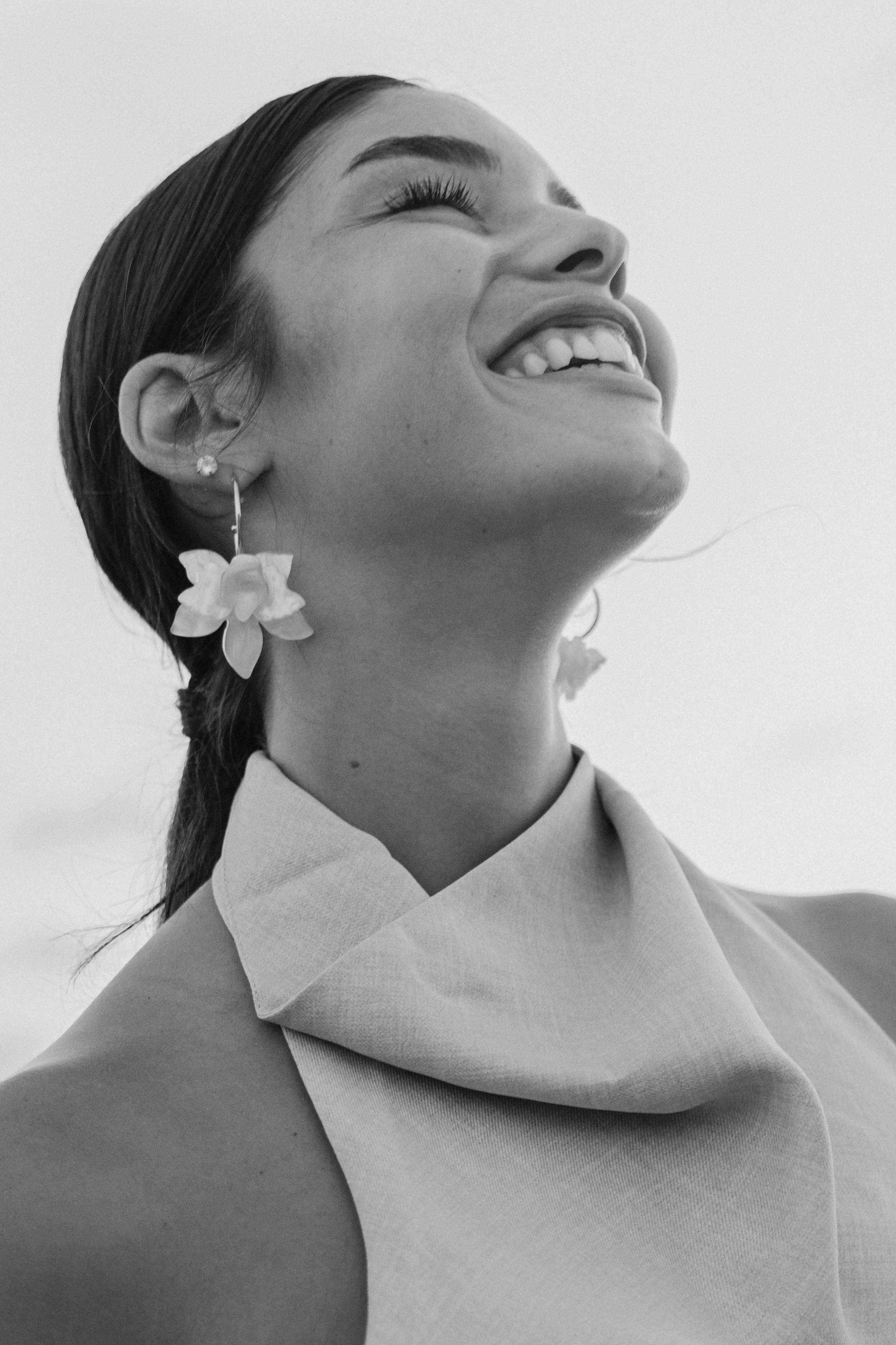 A young model laughing joyfully, with her head tilted back and eyes closed, wearing earrings shaped like flowers and a light-colored top. fashion photography צילומי אופנה למותג