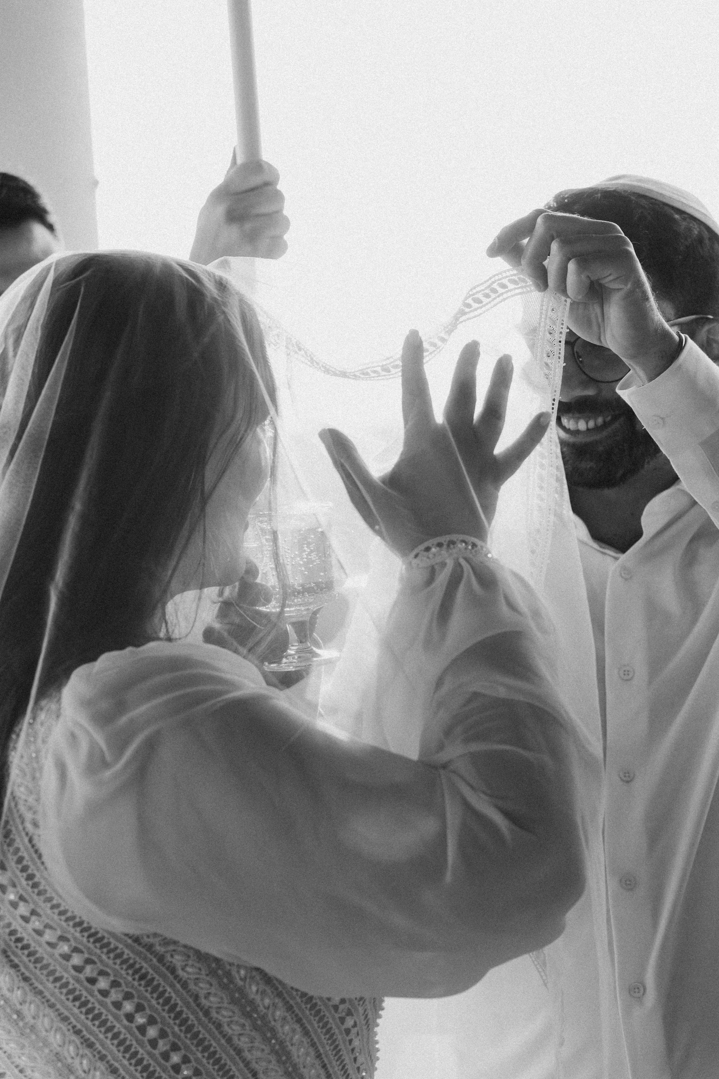 A woman wearing a lace dress and a headscarf is having her hair styled by a man in a white shirt. The man is using a thin lace veil.