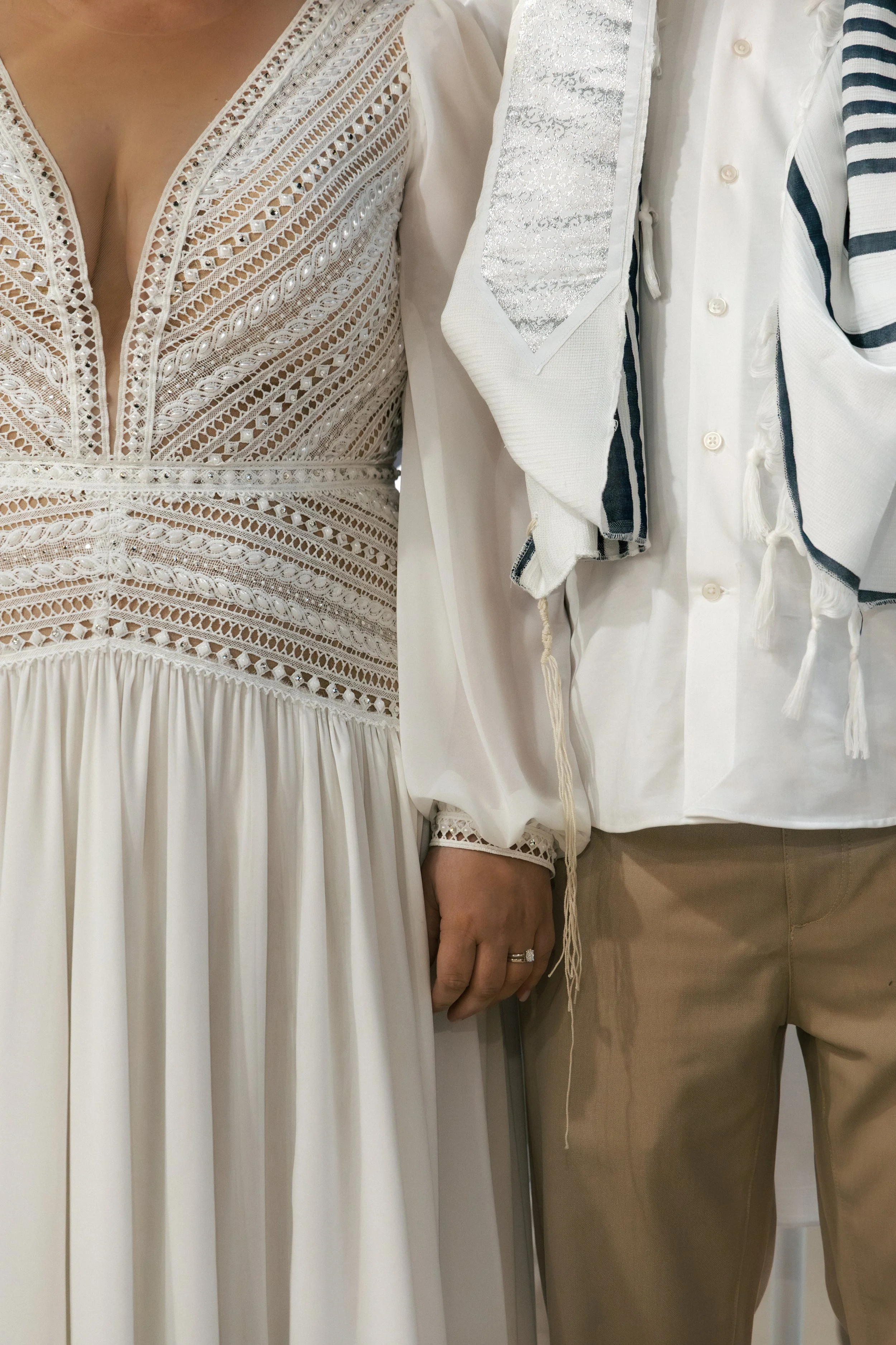 Close-up of an embroidered cream-colored wedding dress and a person dressed in a white shirt with black and white striped fringed sash and khaki pants, standing side by side.