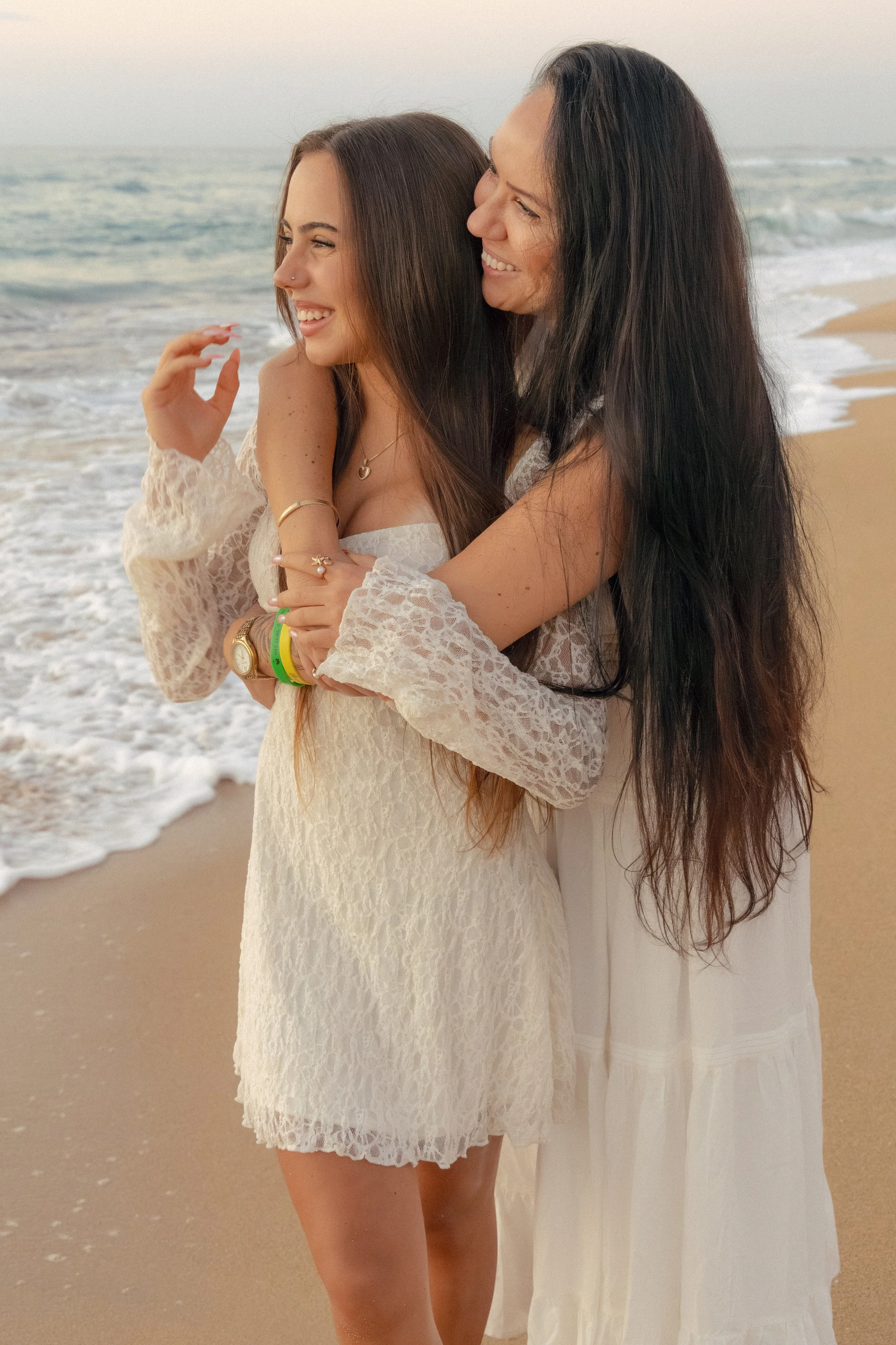 Two women with long dark hair smiling and embracing on the beach near the ocean at sunset, wearing white dresses.