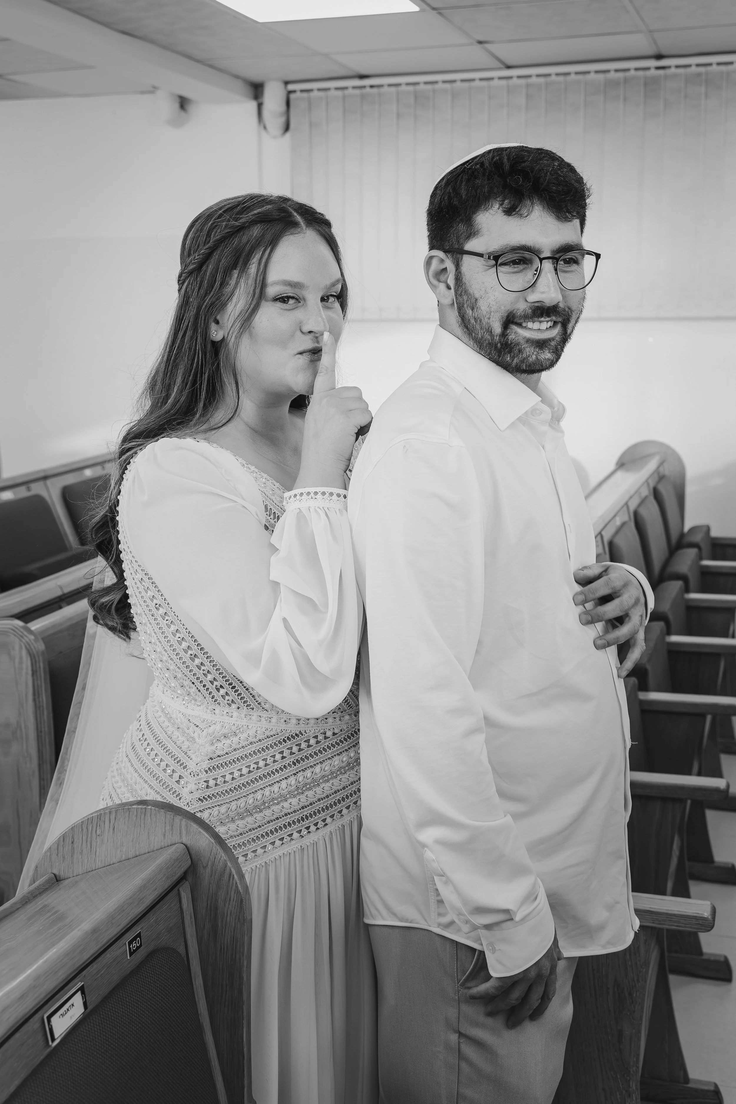 A woman whispering into a man's ear during a religious ceremony or gathering. The woman is making a 'shh' gesture with her finger over her lips, while the man is smiling, wearing glasses, and a kippah. They are standing in a room with wooden pews and