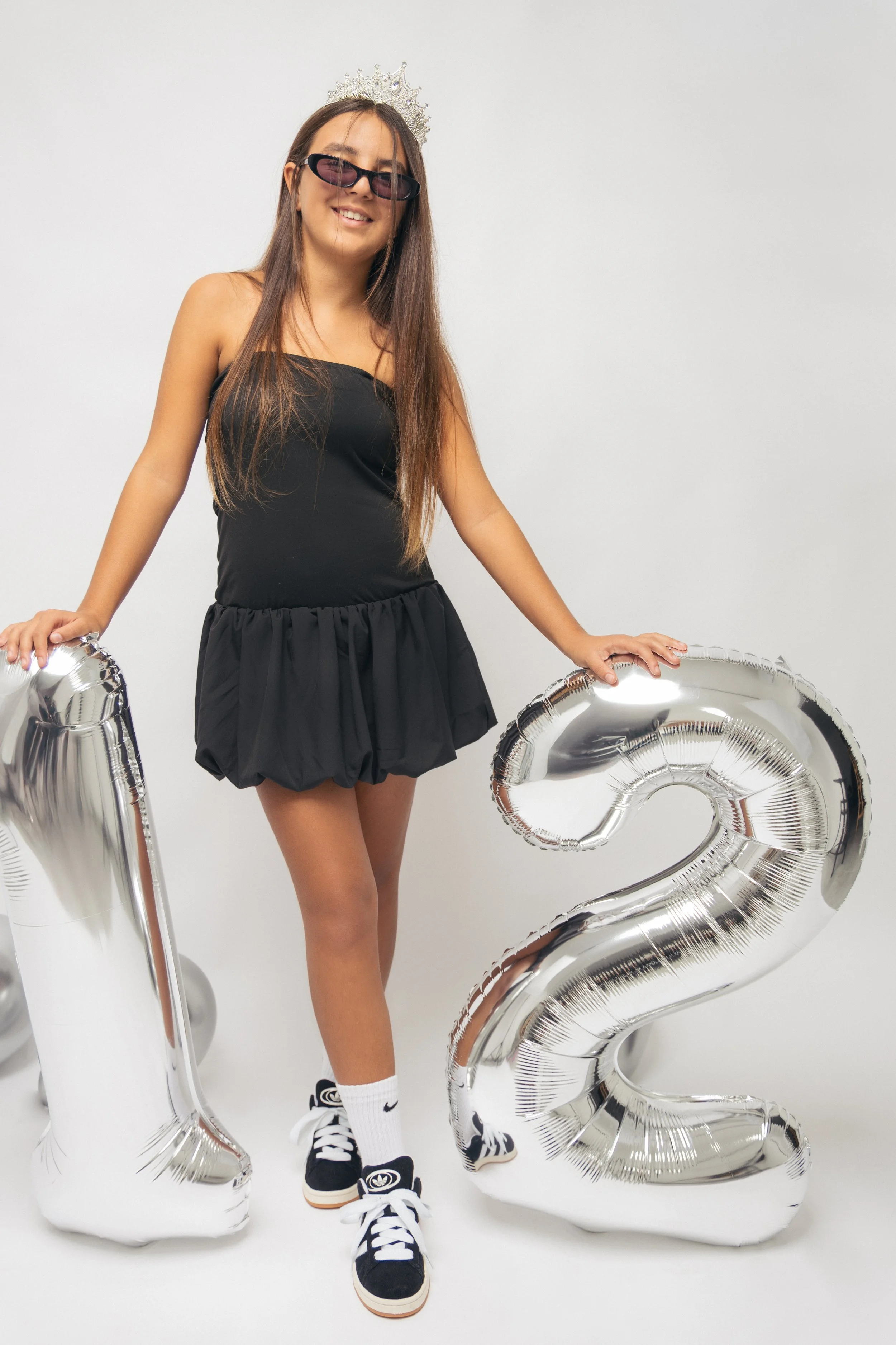 A young woman wearing a black strapless dress, sunglasses, and a tiara, standing against a plain white background, holding large silver balloons in the shape of the number 2 and 3.