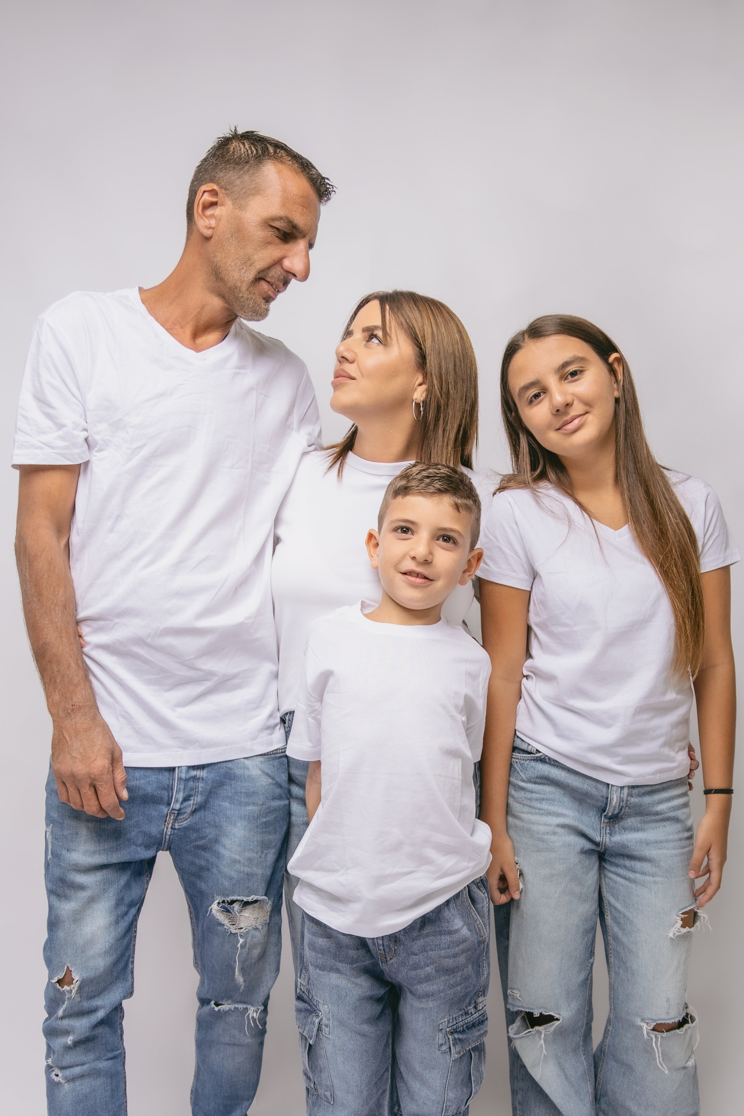 Family of five posing together against a plain gray background, all wearing white T-shirts and distressed jeans, with a mix of smiling and thoughtful expressions.