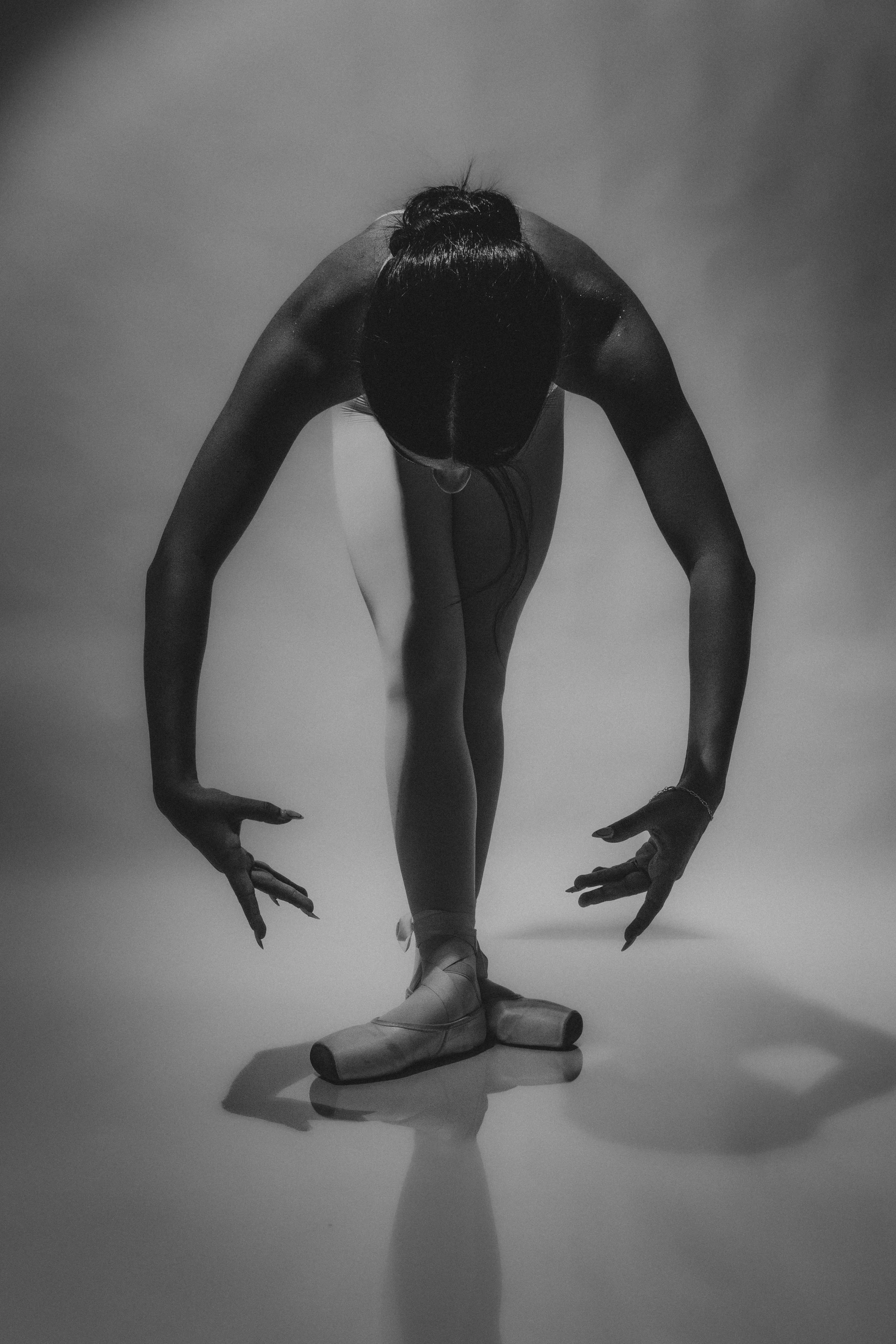 Black and white photo of a ballet dancer in a pose on stage, wearing ballet slippers, with her arms extended downward and head bowed, casting a shadow on the plain background.