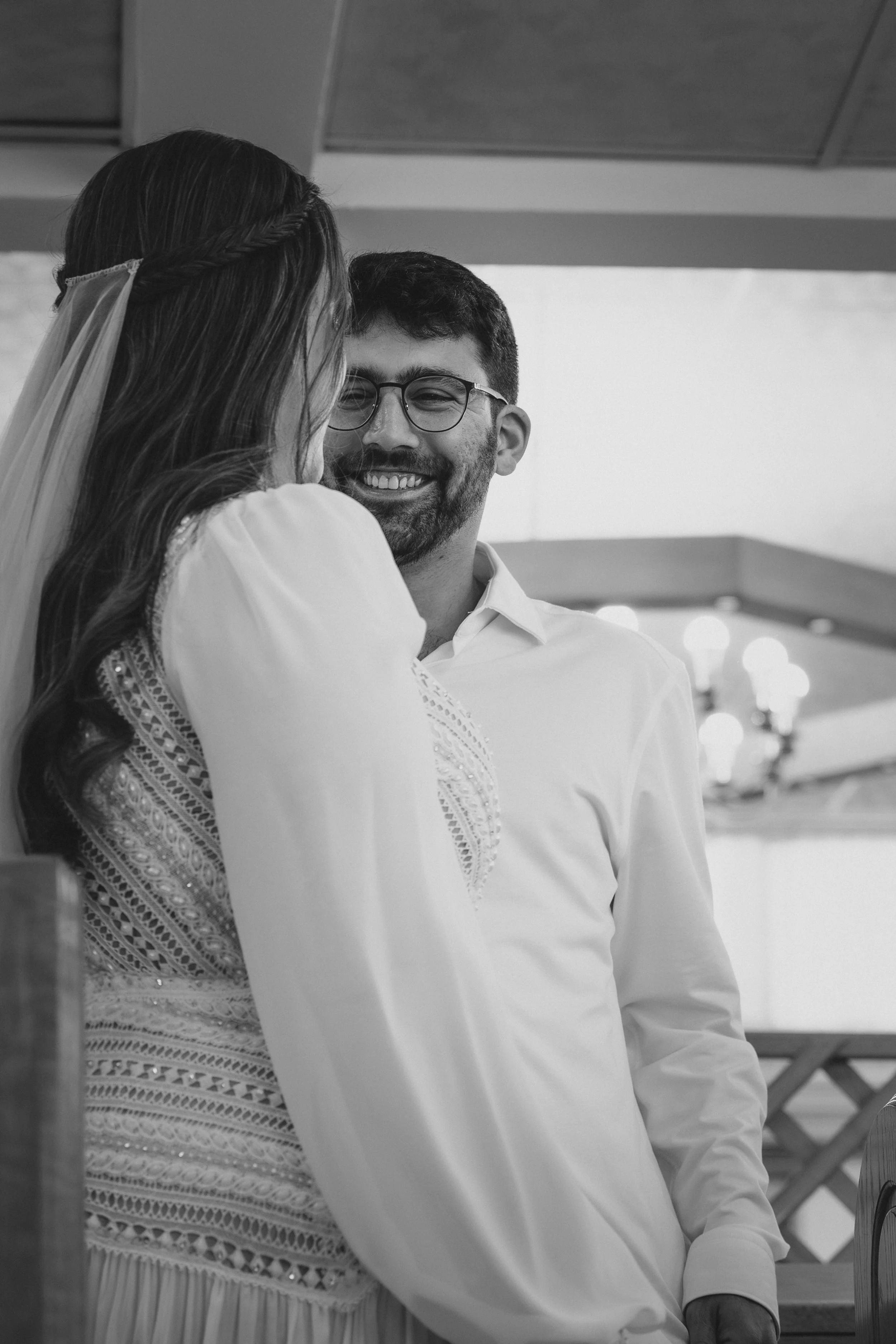 A black and white photo of a smiling man and woman at a close, intimate moment, possibly during a wedding or special event, indoors with ceiling and lighting visible.