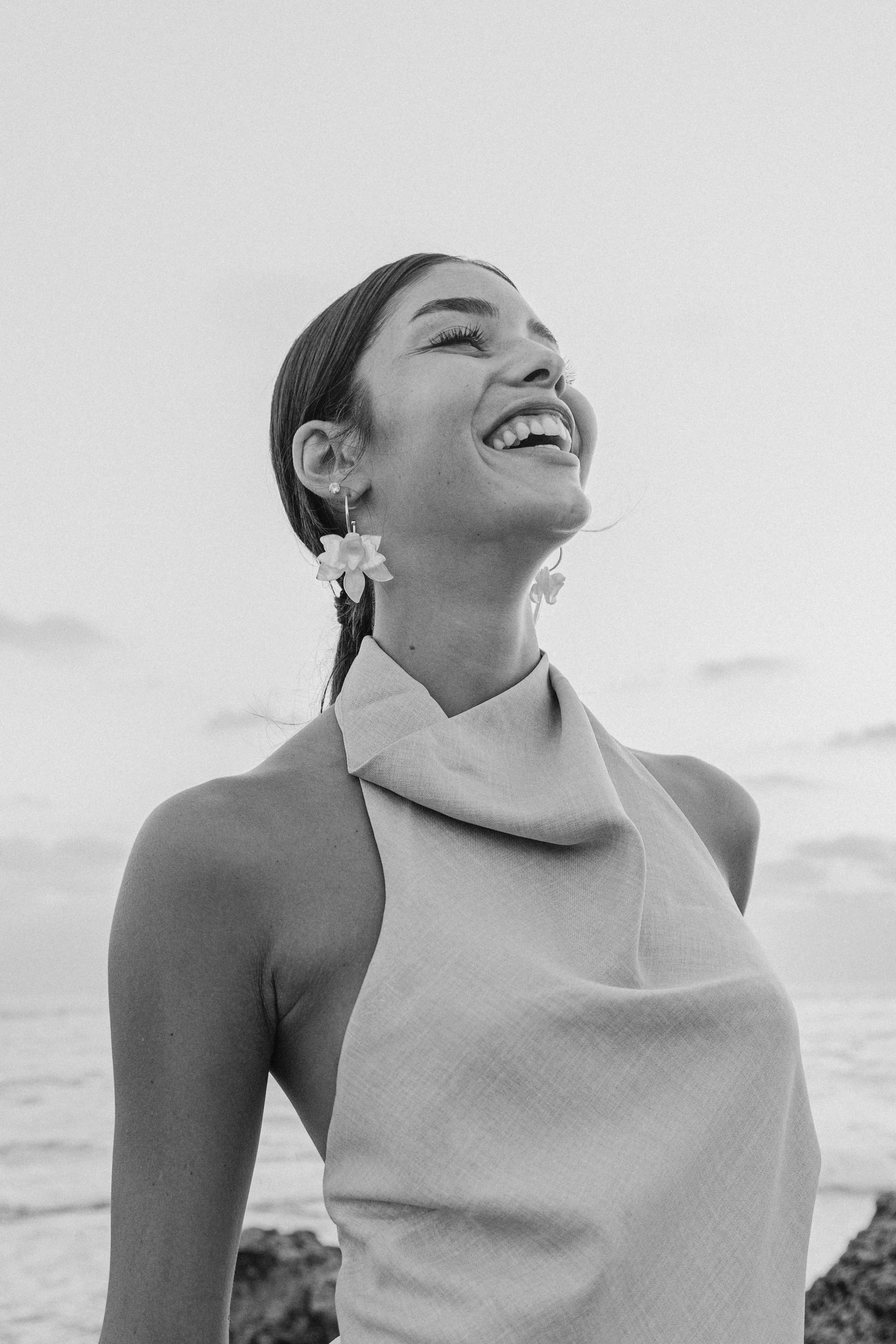 A black and white photo of a woman smiling and looking up, standing outdoors near the ocean with rocks in the background. She is wearing floral earrings and a sleeveless top. fashion photography. צילומי אופנה, צילום למותגים, דוגמנית. פורטרייט