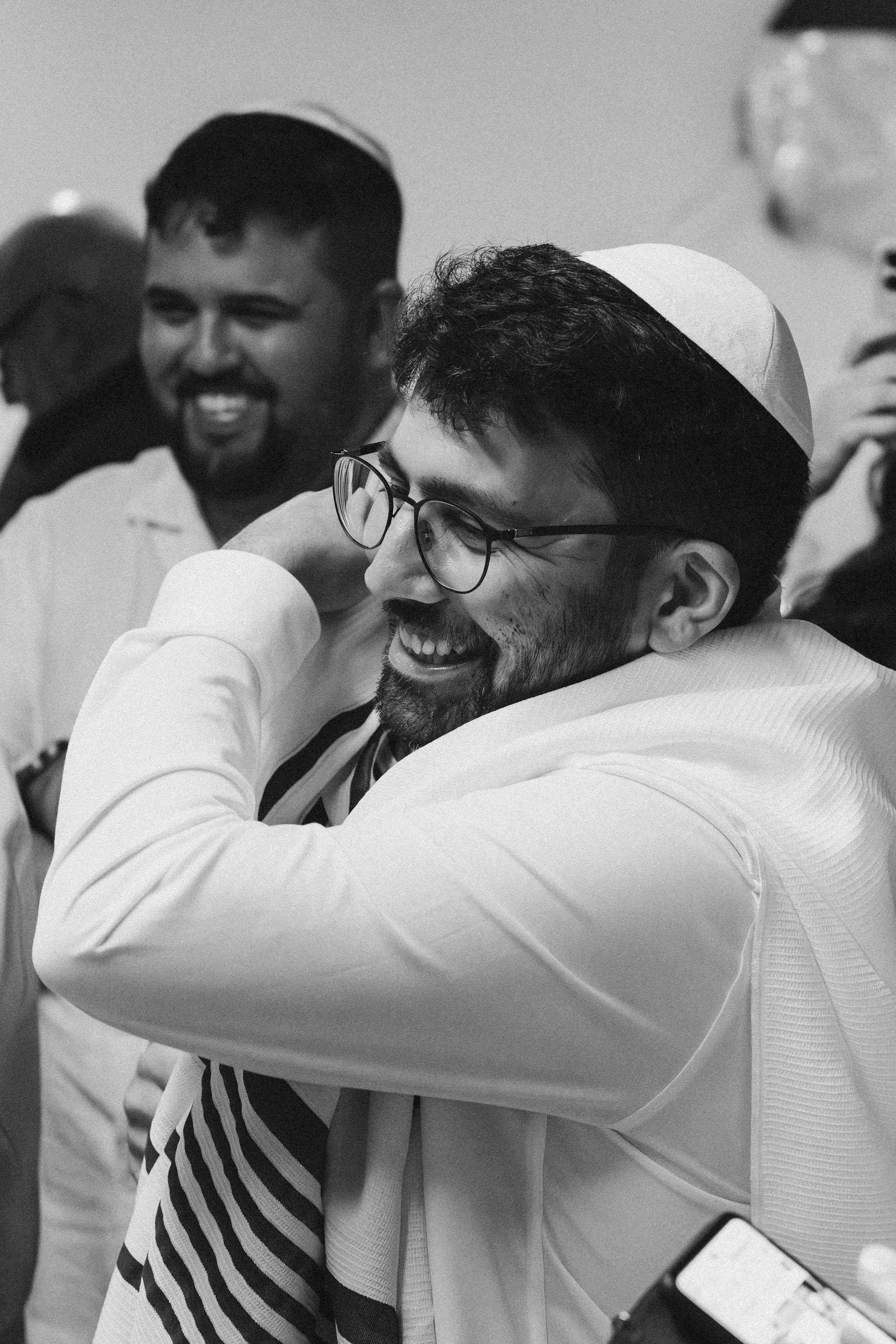 Two men embrace each other and smile, with one wearing glasses and a white kippah, and another man in the background smiling.