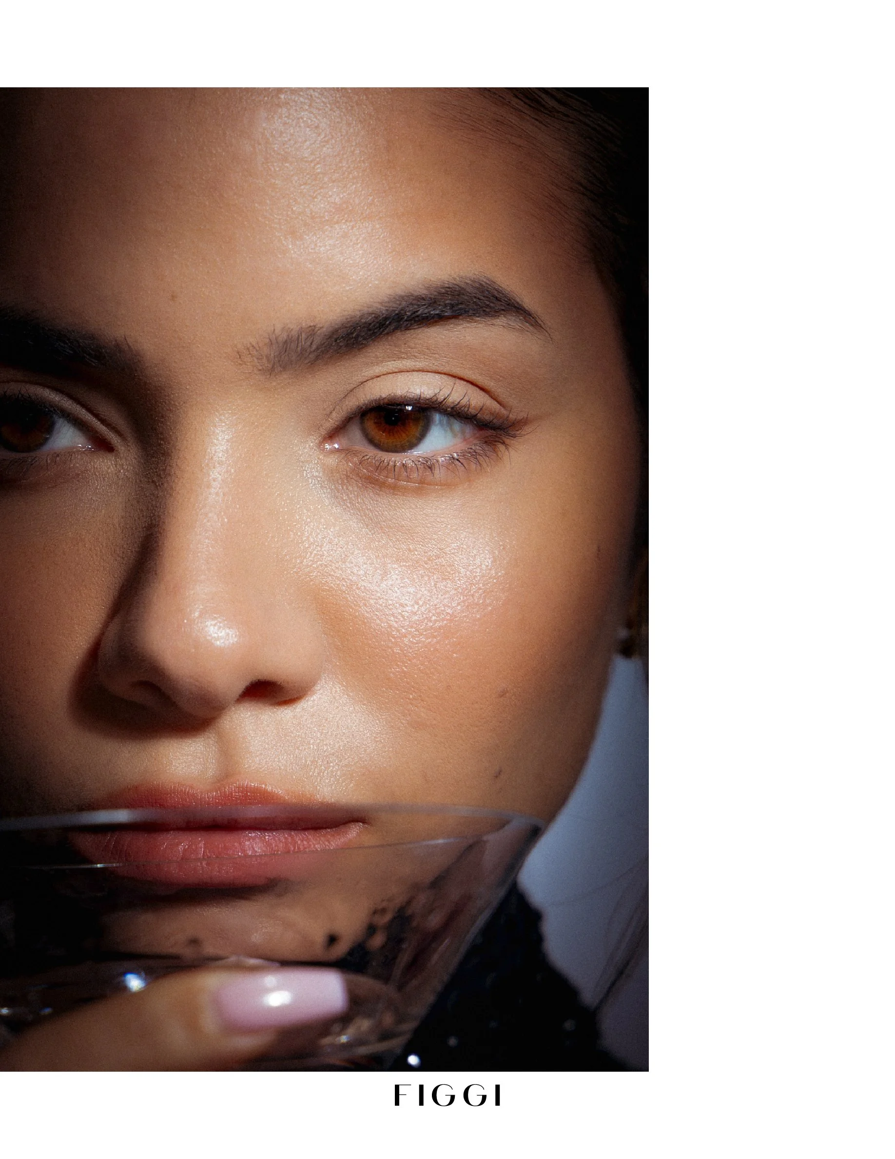 Close-up of a woman with brown eyes, dark eyebrows, and smooth skin, holding a glass of beverage near her chin, with the word 'FIGGI' written at the bottom.