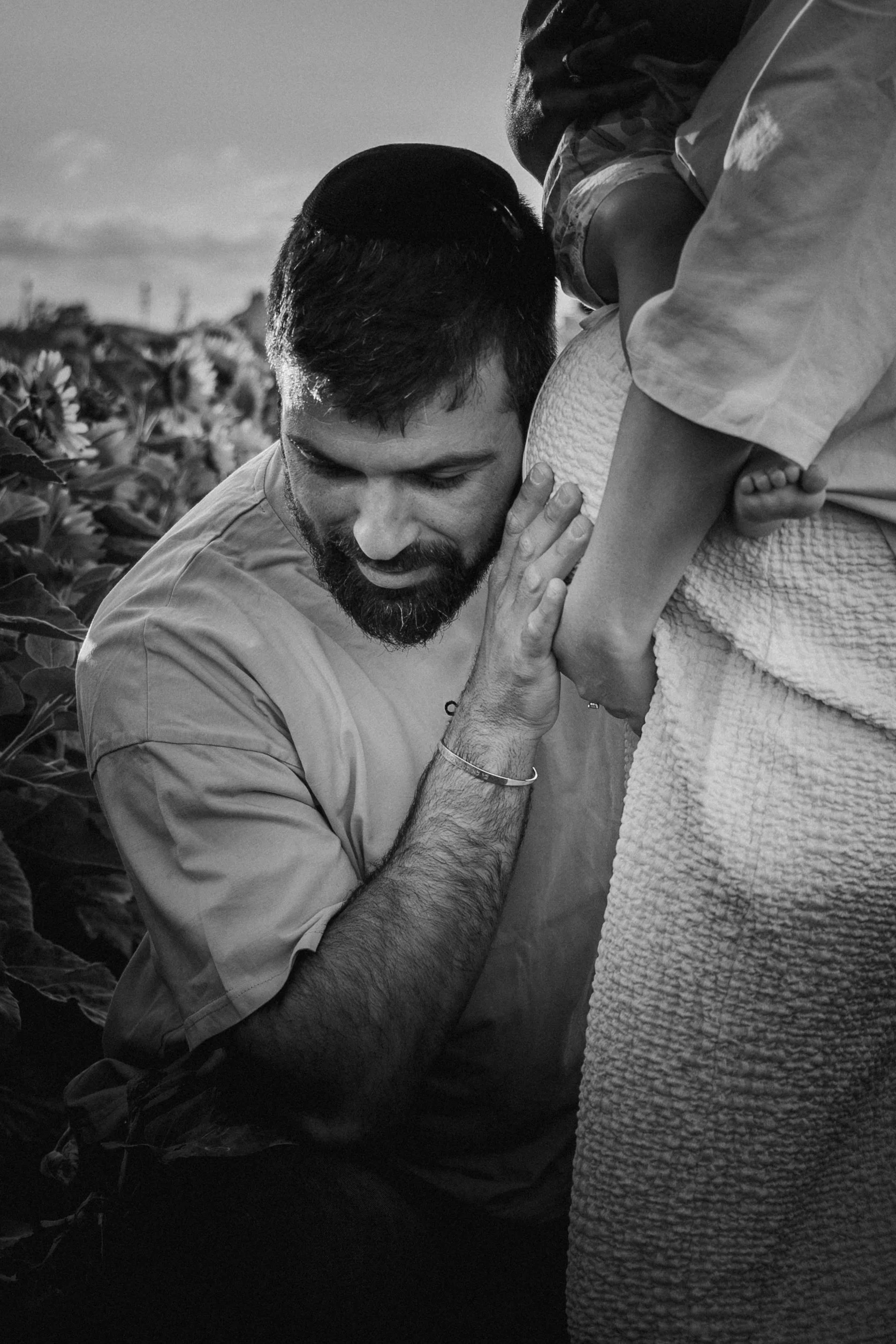A man holding a child close to his chest in an outdoor setting, with a field of sunflowers in the background. The photo is in black and white.