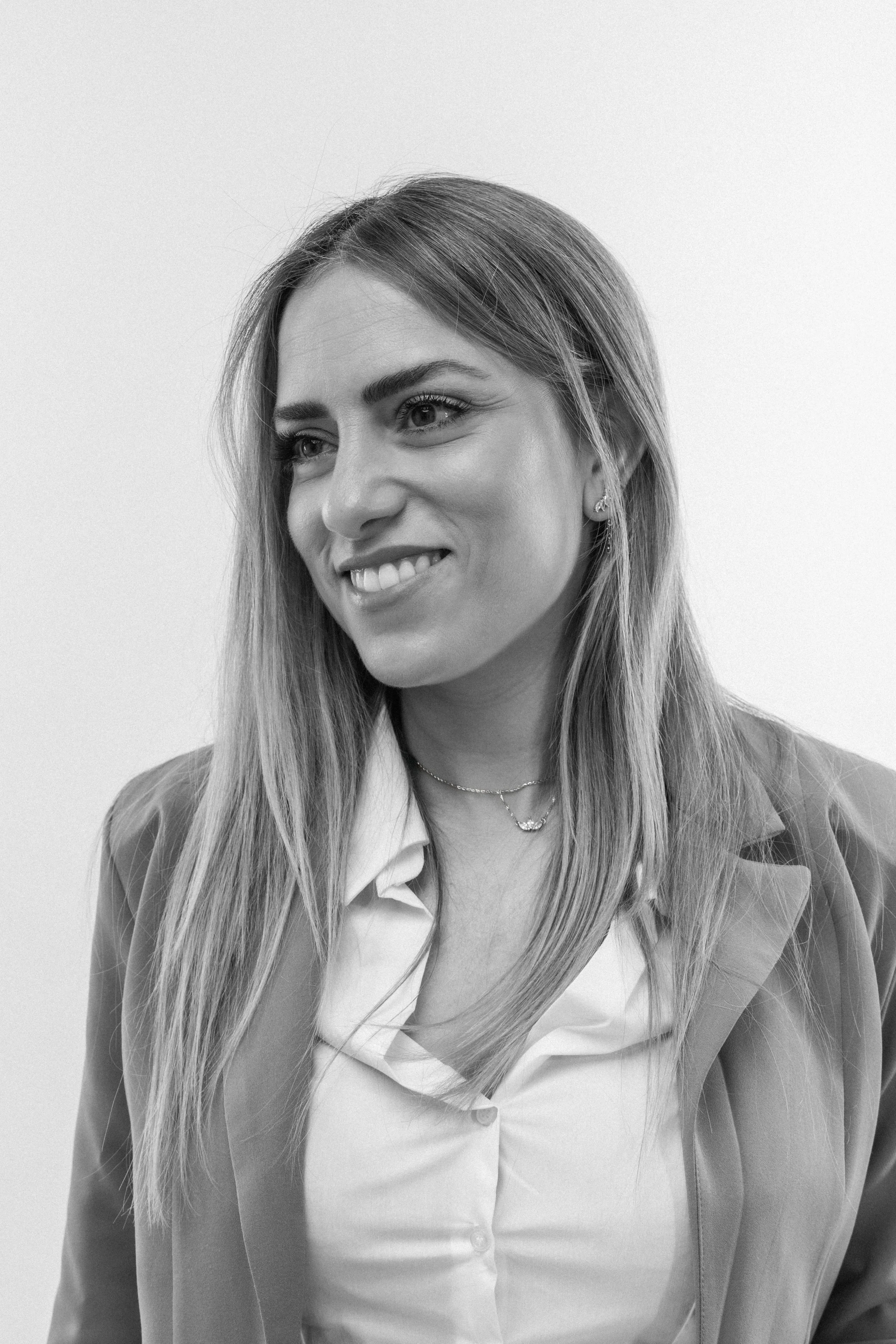 Black and white portrait of a young woman with long hair, smiling, wearing a blazer, shirt, necklace, and earrings.