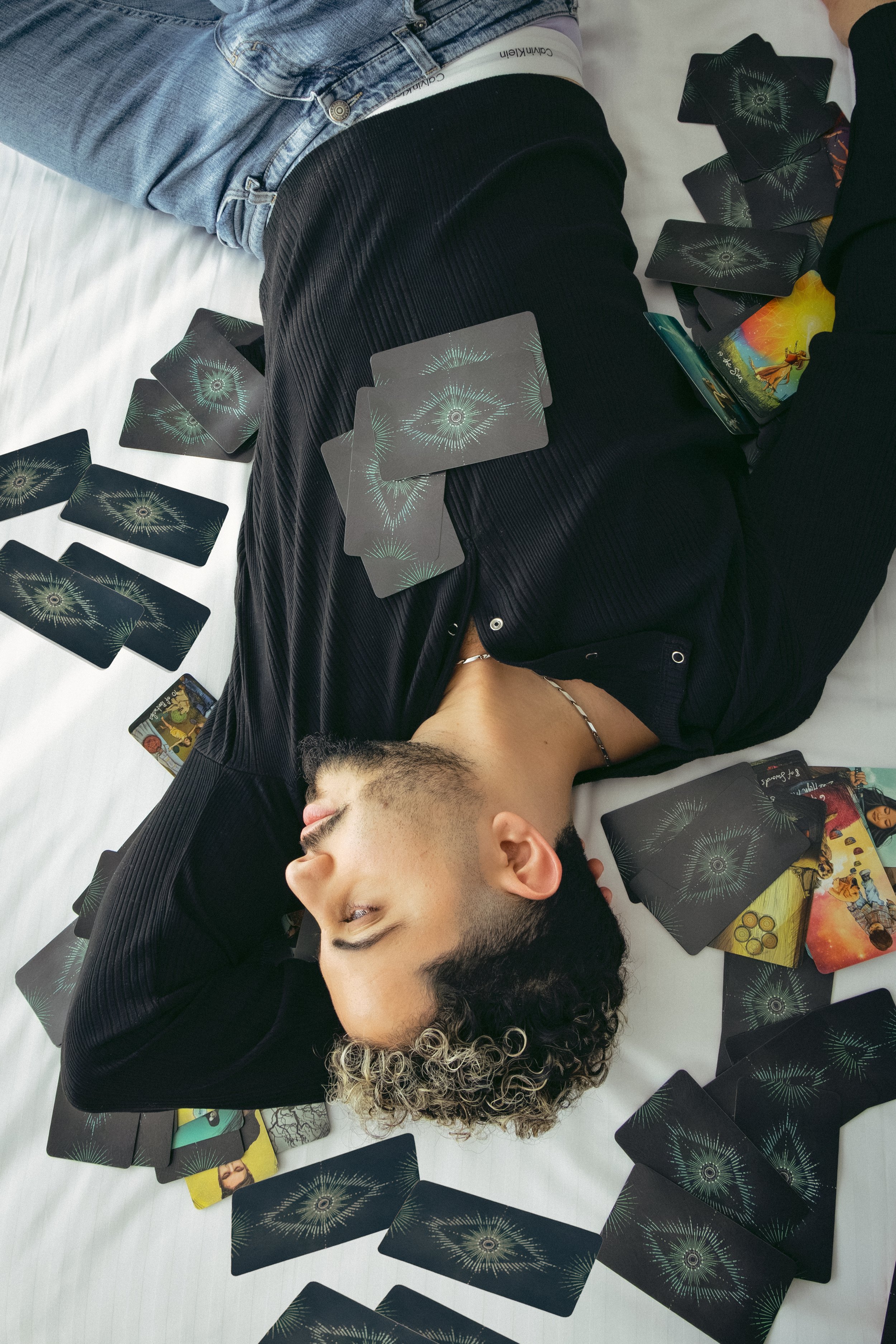 A person with curly hair and a black shirt lying on a bed surrounded by tarot cards