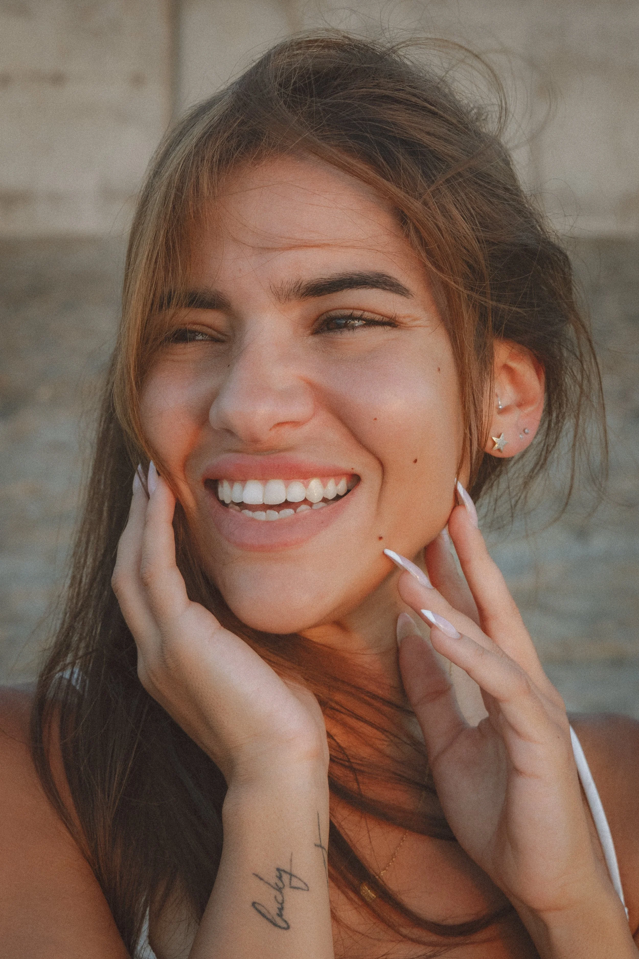 A young woman with long brown hair smiling while holding her face with both hands. She has multiple earrings on her left ear, including a star-shaped earring, and wears a delicate gold necklace. The background is blurry.