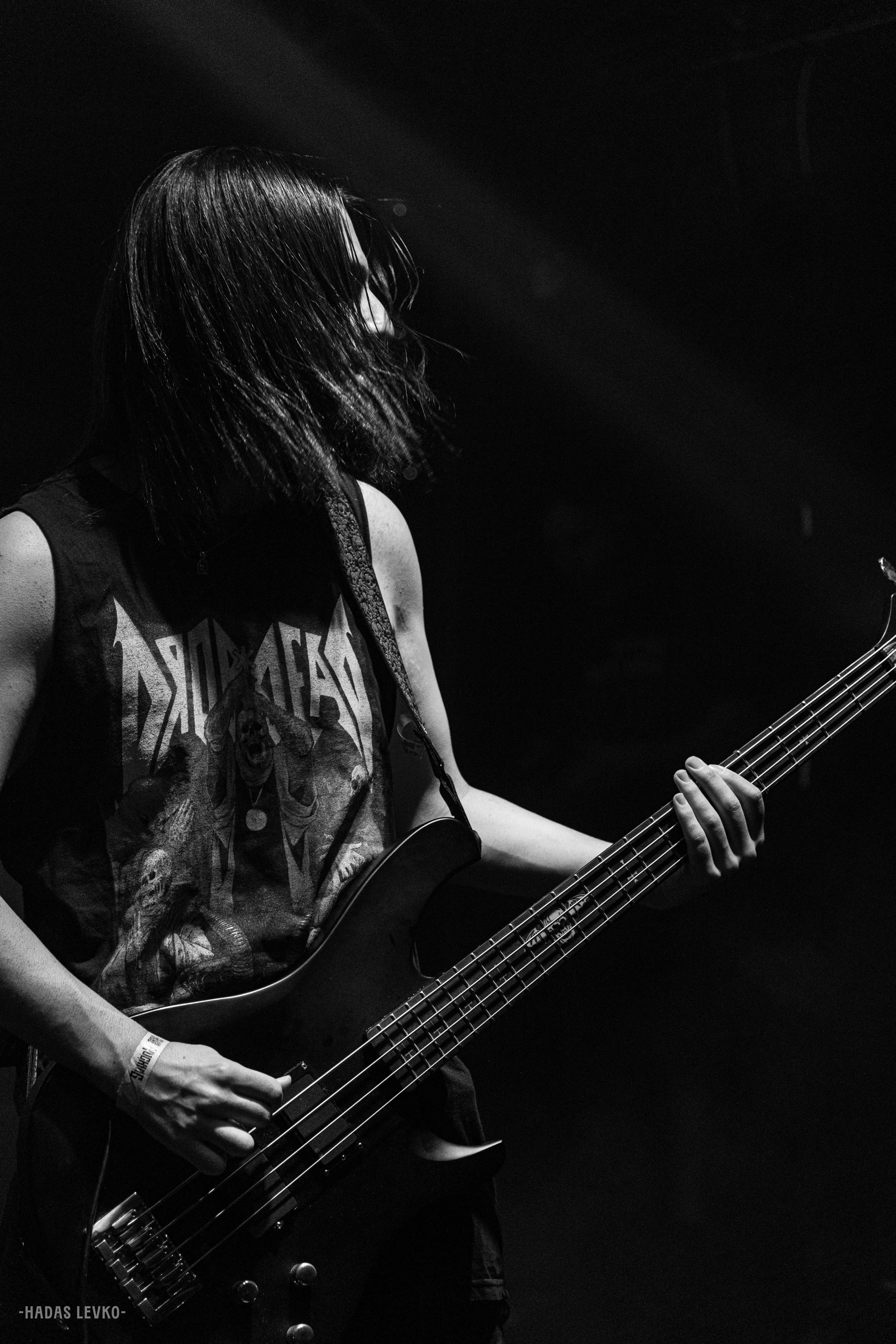 A person with long hair playing a bass guitar in a dark setting, wearing a sleeveless shirt with a graphic design.